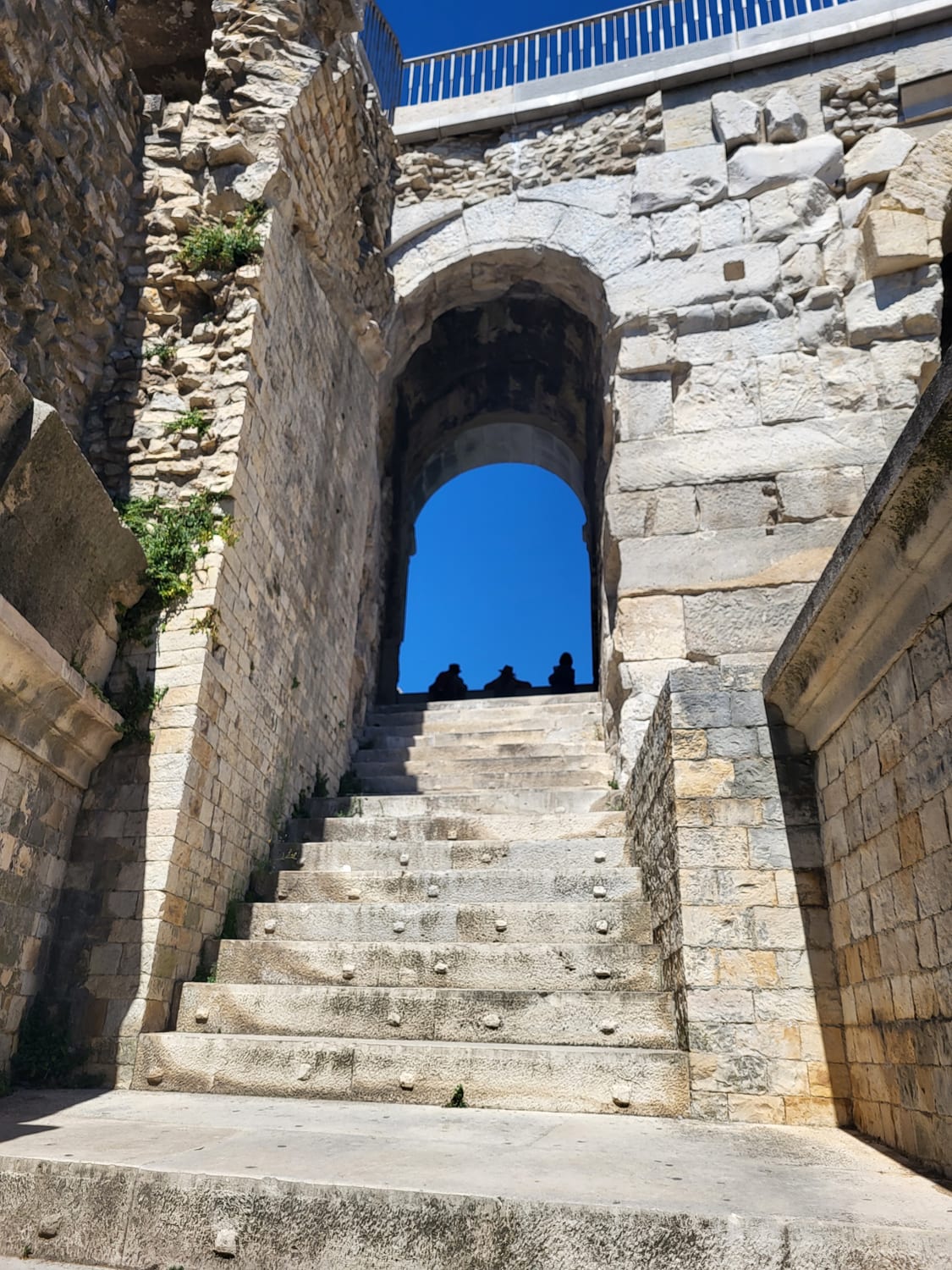 una escalera de piedra de la Arena de Nimes que sube hacia el arco de un vomitorio, con las siluetas de algunos visitantes en las gradas
