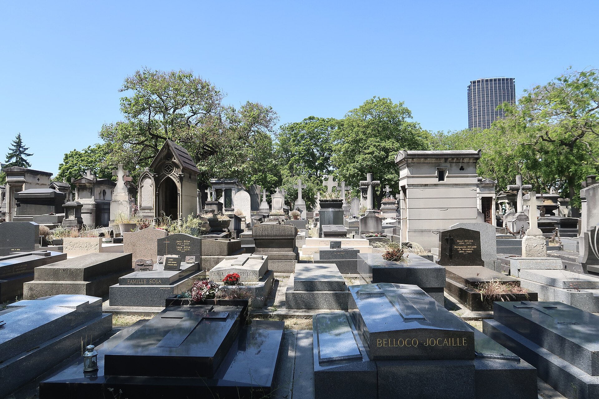 il cimitero di montparnasse a parigi, con le lastre di granito scuro in primo piano, le cappelle di famiglia e una tomba neogotica col tetto a punta in mezzo agli alberi