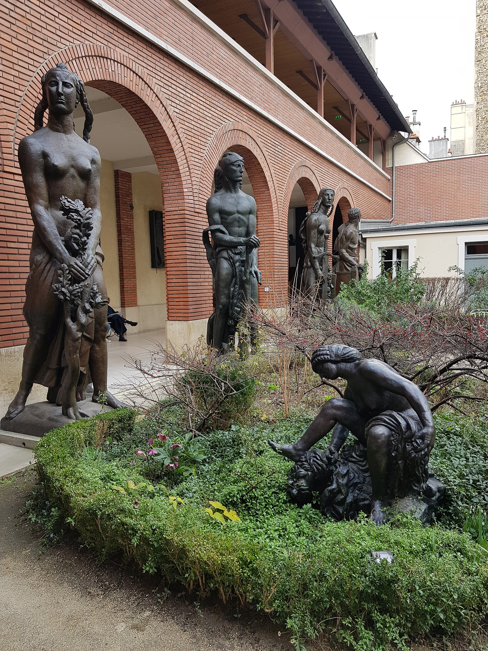 il giardino del musee bourdelle a parigi, con i grandi bronzi in piedi sotto le arcate di mattone rosso del portico e una figura accovacciata in mezzo alle siepi basse