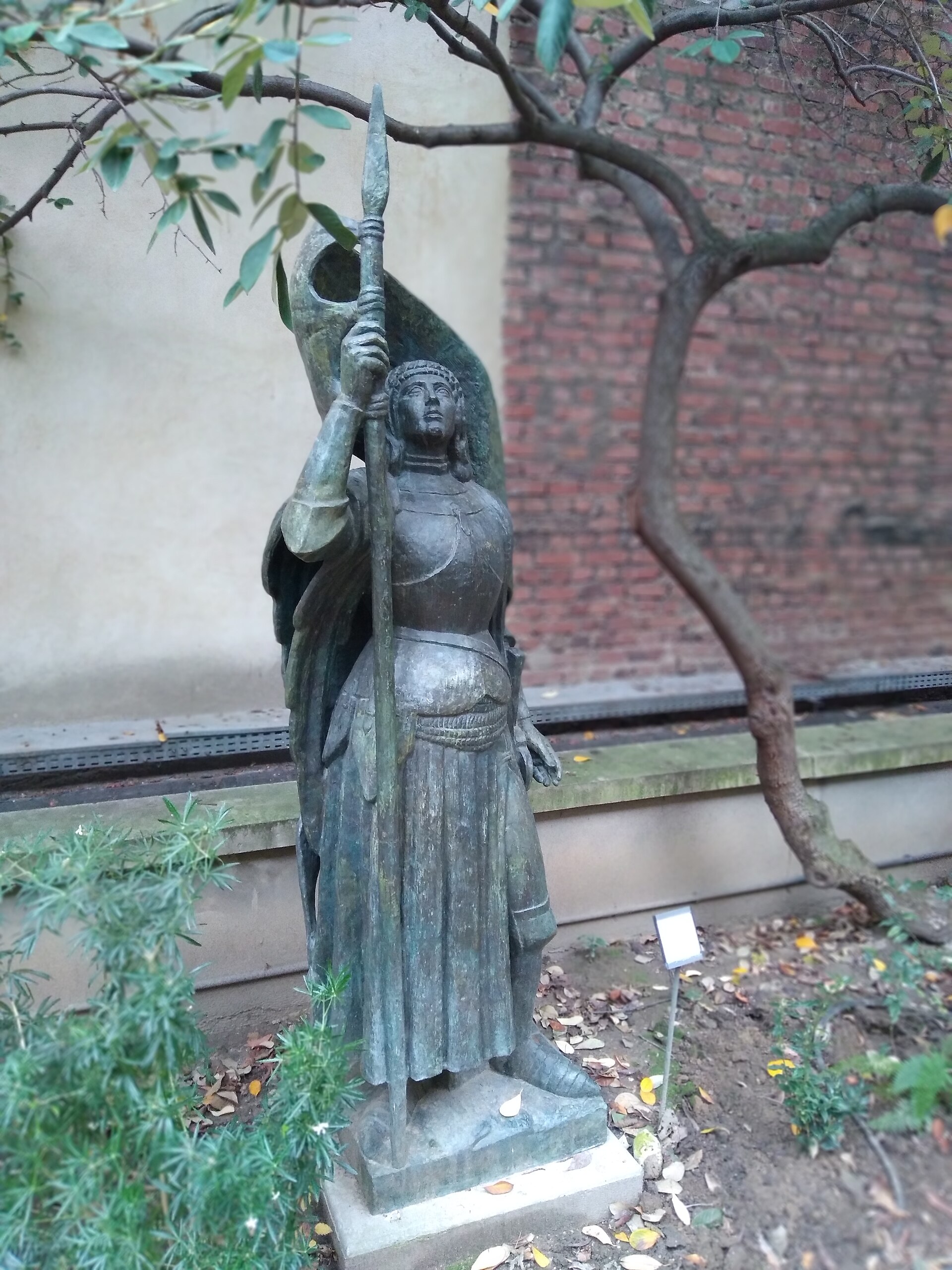 la statua in bronzo di giovanna d arco di antoine bourdelle nel giardino del museo, in piedi su un basso zoccolo di pietra, con la lancia alzata nella mano destra e la patina verde, davanti a un muro di mattoni e a un ramo spoglio