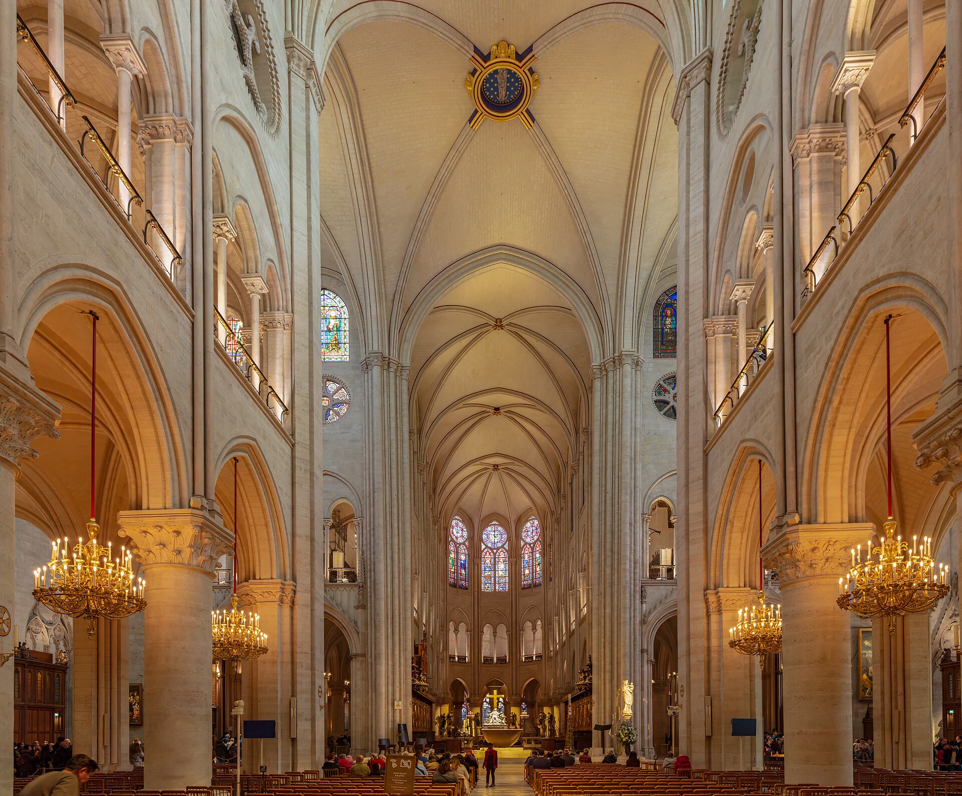 la navata interna di notre-dame restaurata, con le alte volte gotiche chiare, i lampadari dorati accesi lungo le colonne e i fedeli seduti nei banchi verso l’altare