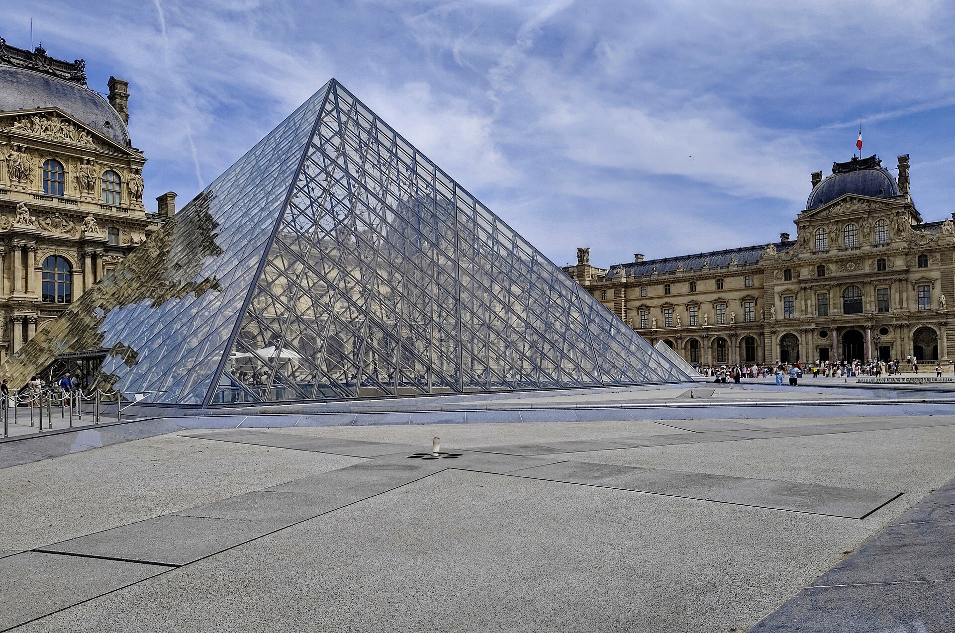 la piramide di vetro del louvre nella cour napoleon, con i palazzi ottocenteschi ai due lati e i visitatori raccolti sul piazzale davanti all’ingresso