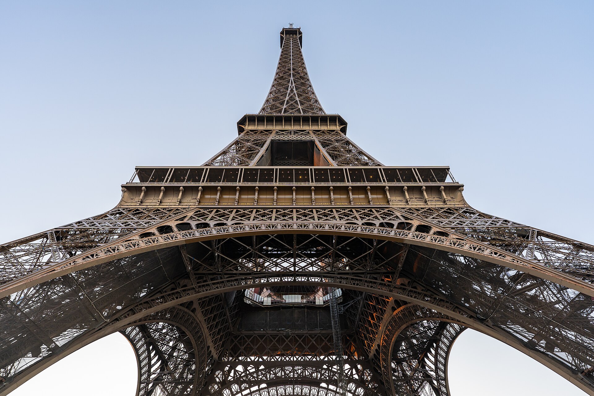 la torre eiffel di parigi vista da sotto, con la struttura reticolare di ferro che si restringe verso il cielo azzurro e le travi che si intrecciano a raggiera sopra la testa
