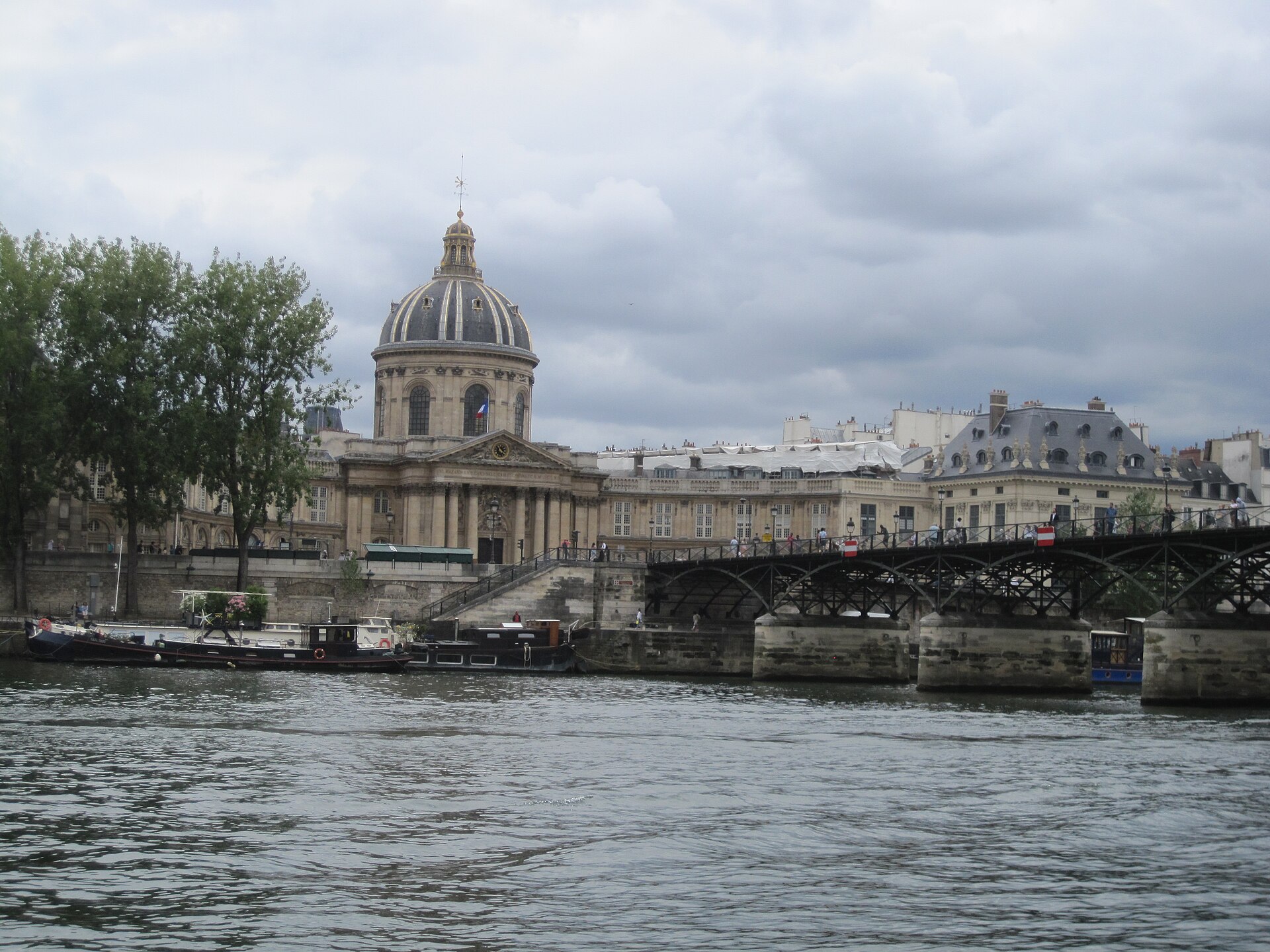 l institut de france visto dalla senna a parigi, con la cupola scura sopra il colonnato, le arcate metalliche del pont des arts a destra e le chiatte ormeggiate lungo il quai