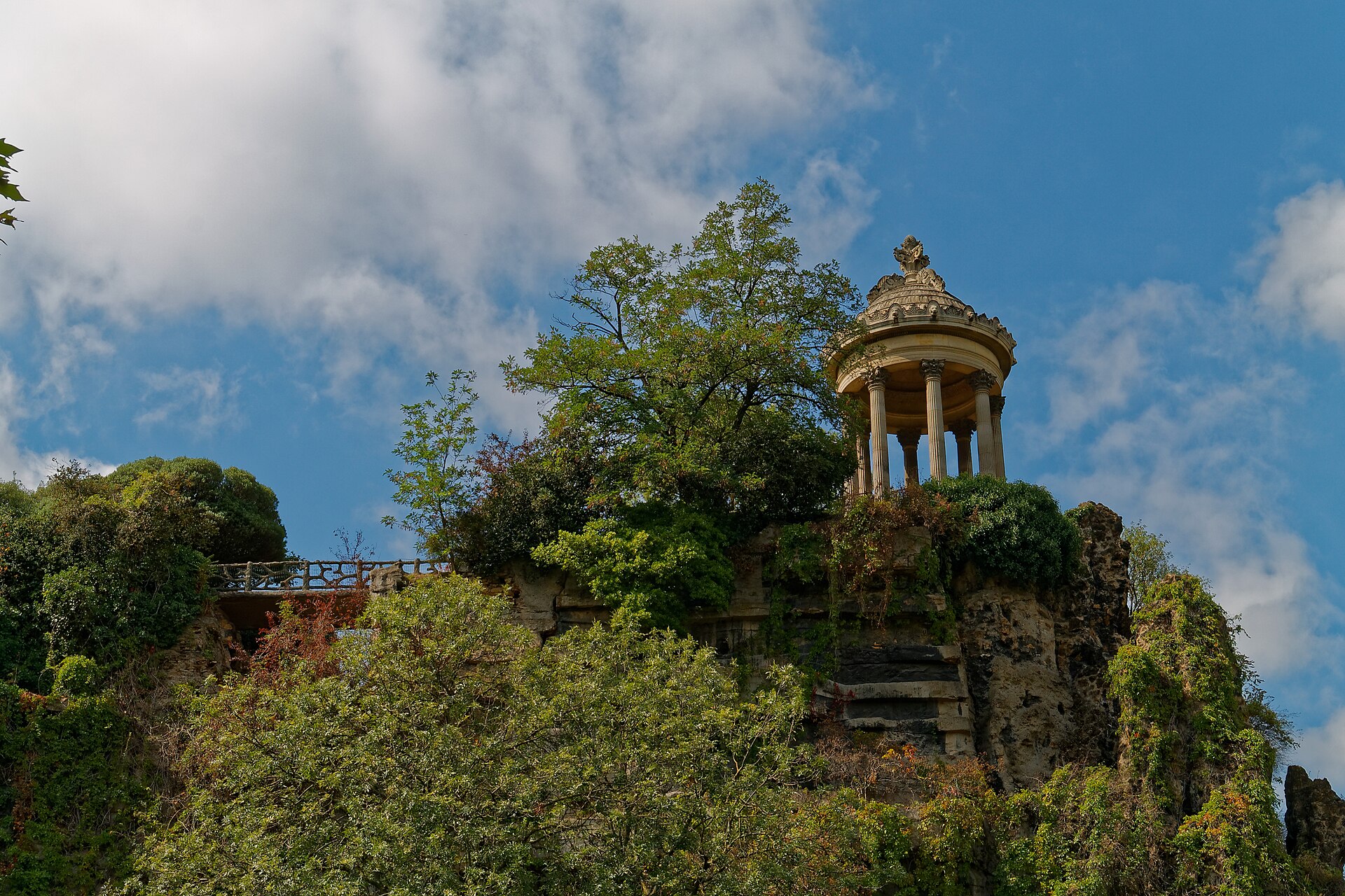 il temple de la sibylle nel parco delle buttes chaumont a parigi, il tempietto circolare a colonne con la cupoletta in cima alla rupe coperta di vegetazione, sotto un cielo con nuvole bianche