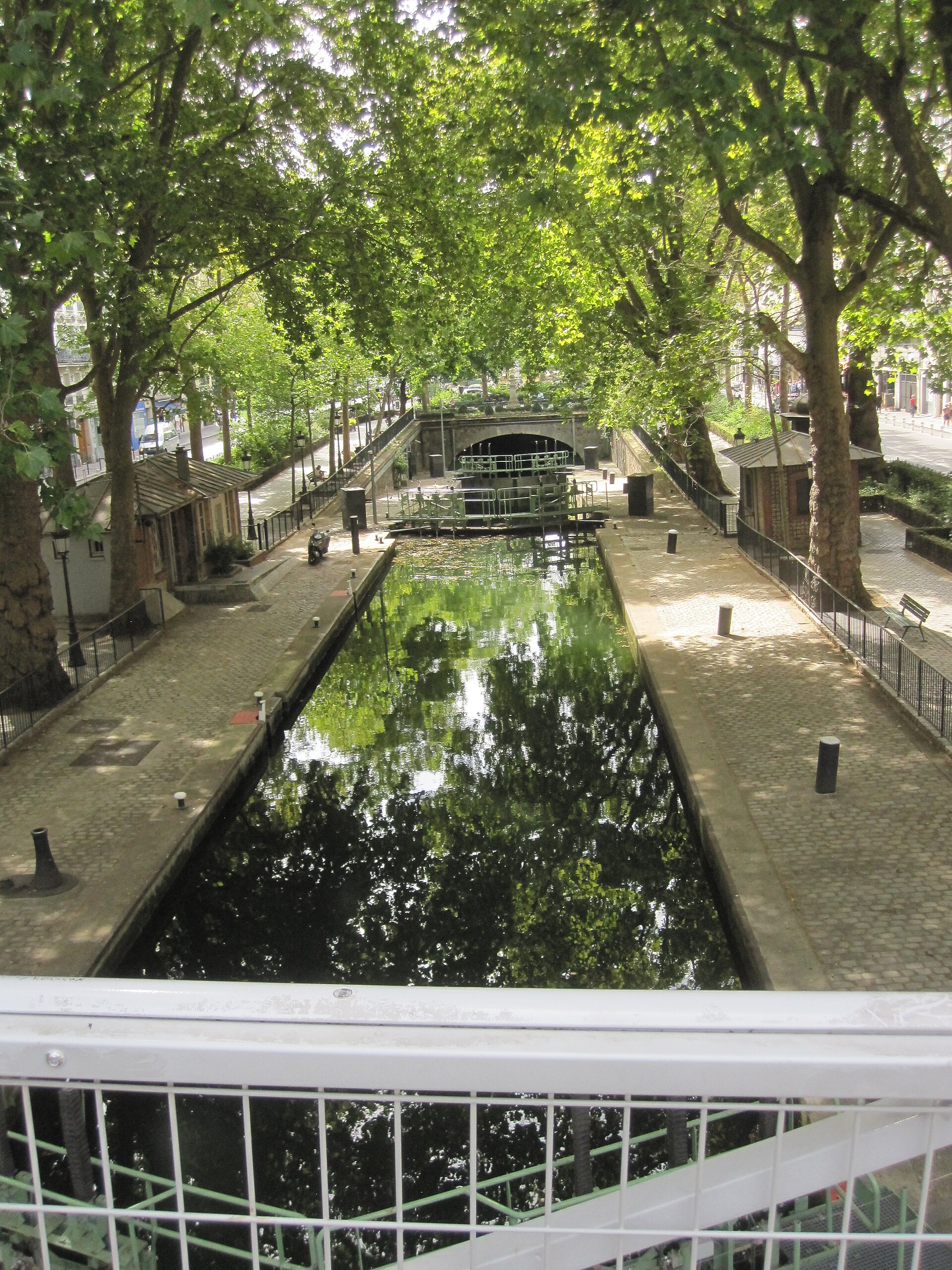 una chiusa del canal saint martin a parigi vista dall alto di una passerella, l acqua scura che riflette i platani, i due casotti di servizio sulle banchine lastricate e la volta del passaggio coperto in fondo