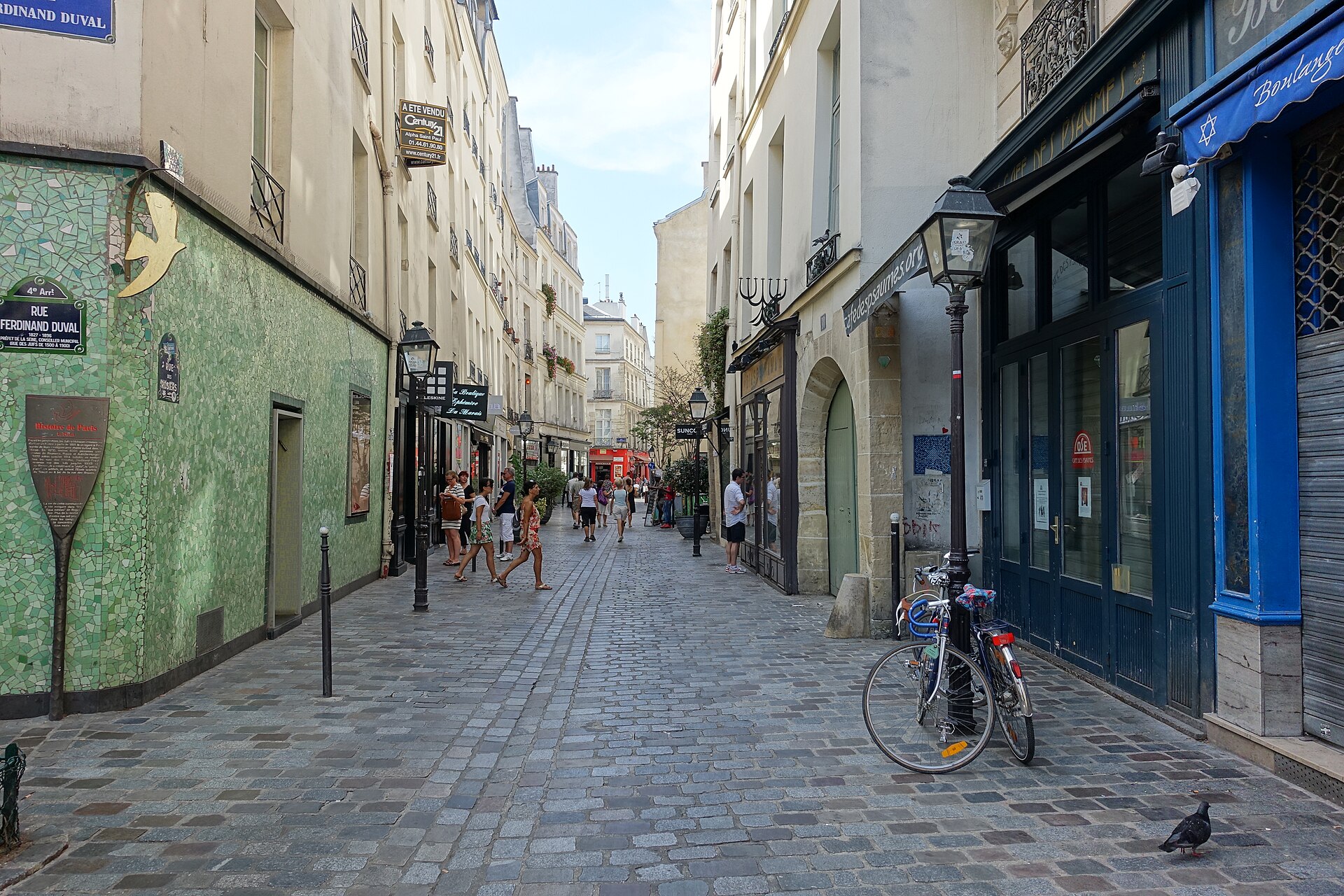 la rue des rosiers nel quartiere ebraico del marais a parigi, con il lastricato pedonale, le insegne dei negozi, un muro rivestito di mosaico verde e una bicicletta appoggiata a un lampione