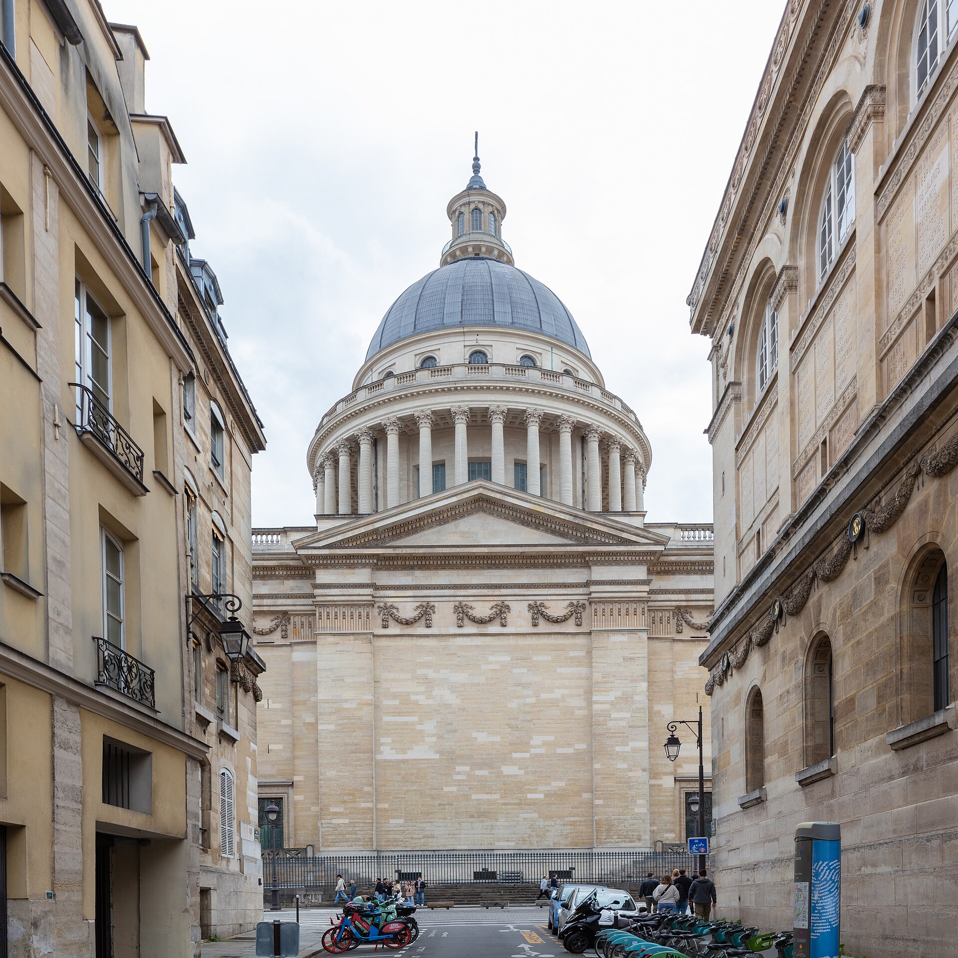 la cupola e il colonnato del pantheon di parigi visti in fondo a una via stretta, con la facciata della biblioteca sainte genevieve sulla destra e le biciclette parcheggiate lungo la strada