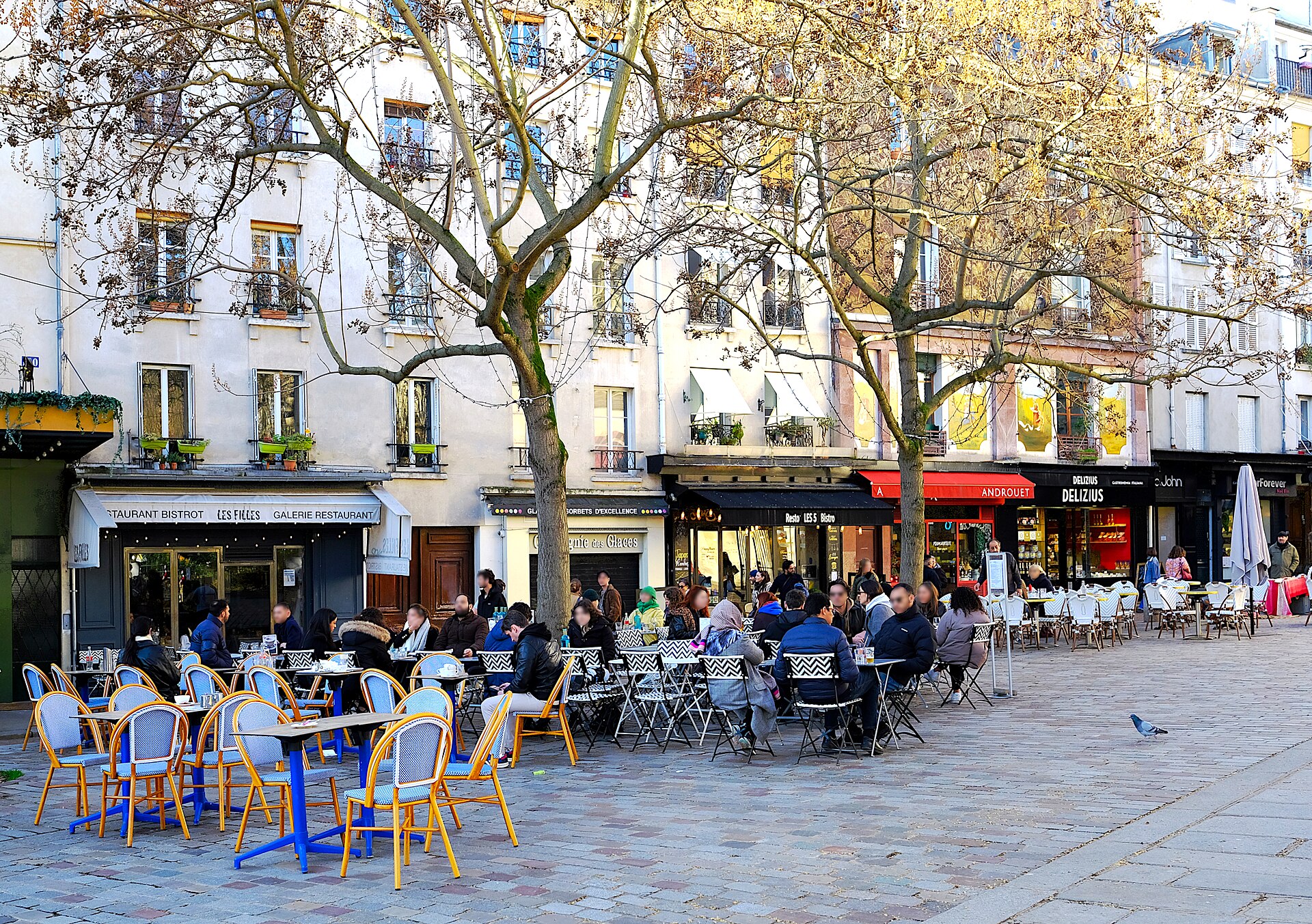 le terrazze dei caffe di rue mouffetard a parigi sotto i platani spogli, con le sedie da bistrot gialle e blu, i tavolini occupati e le insegne dei negozi sul fondo