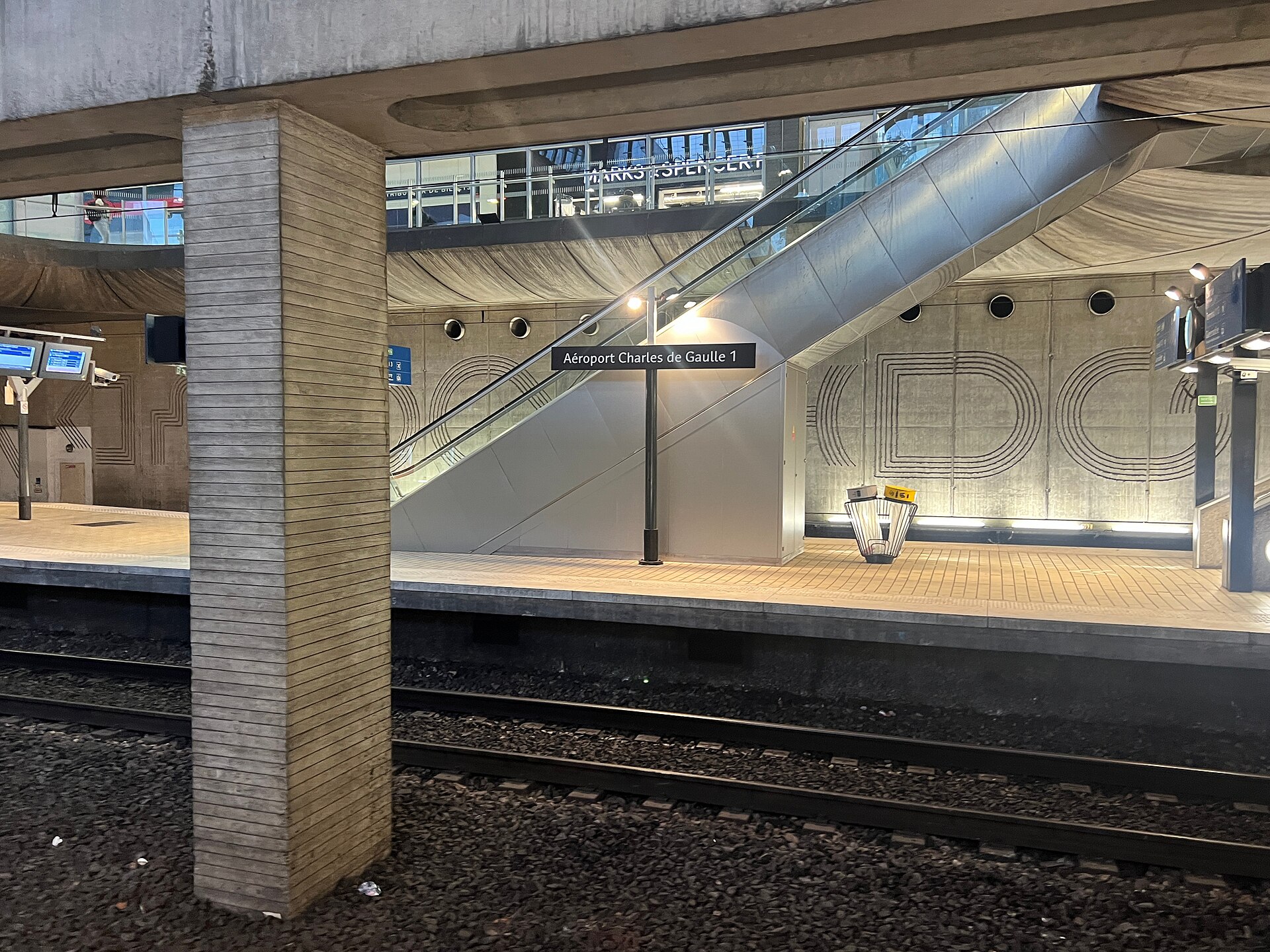 la stazione rer aeroporto charles de gaulle 1 con la scala mobile, il cartello blu del nome della fermata e i manifesti pubblicitari gialli