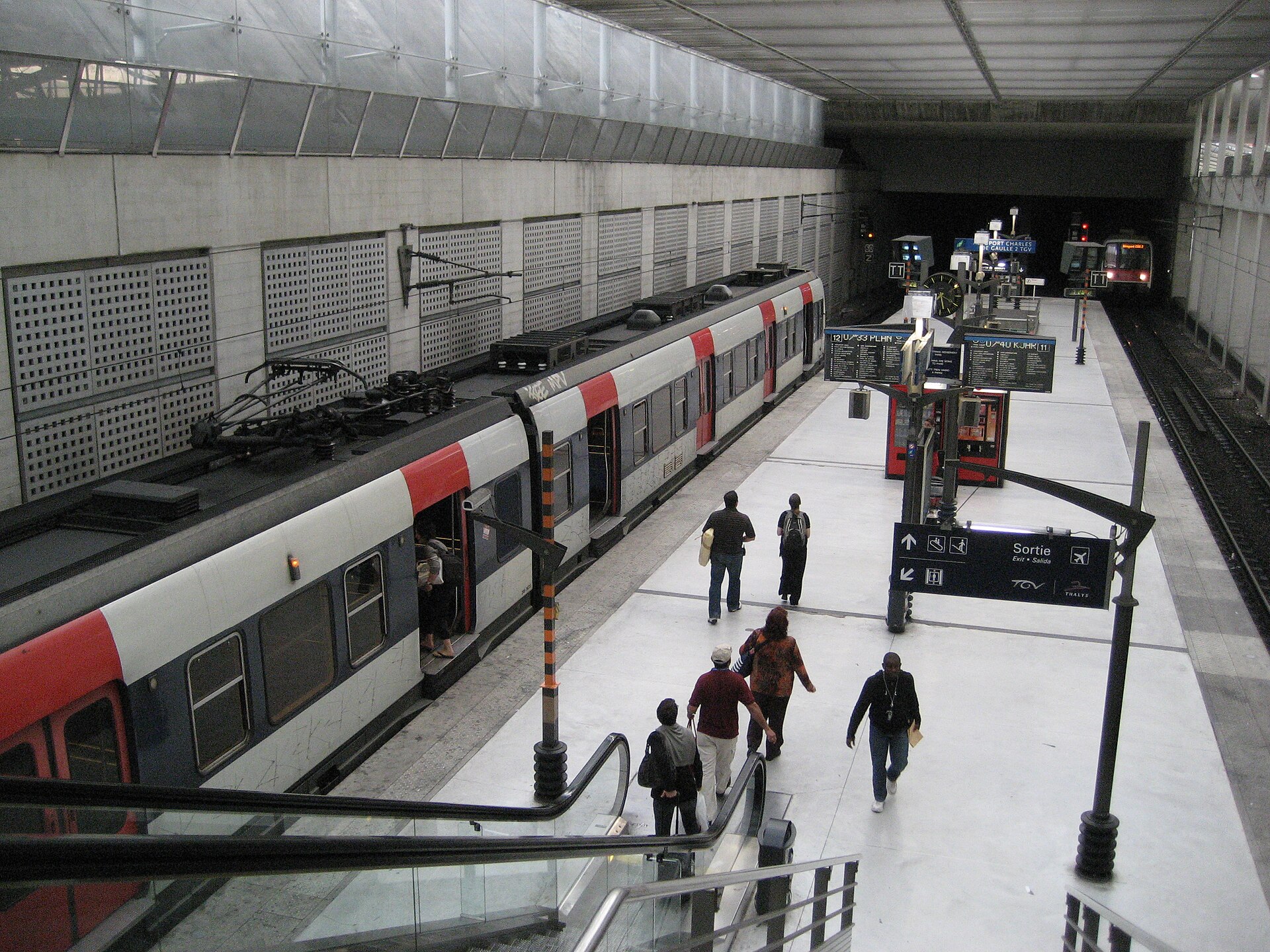 un treno rer b fermo al binario della stazione aeroporto charles de gaulle 1, con la scala mobile in primo piano e il cartello sortie con le icone tgv e thalys