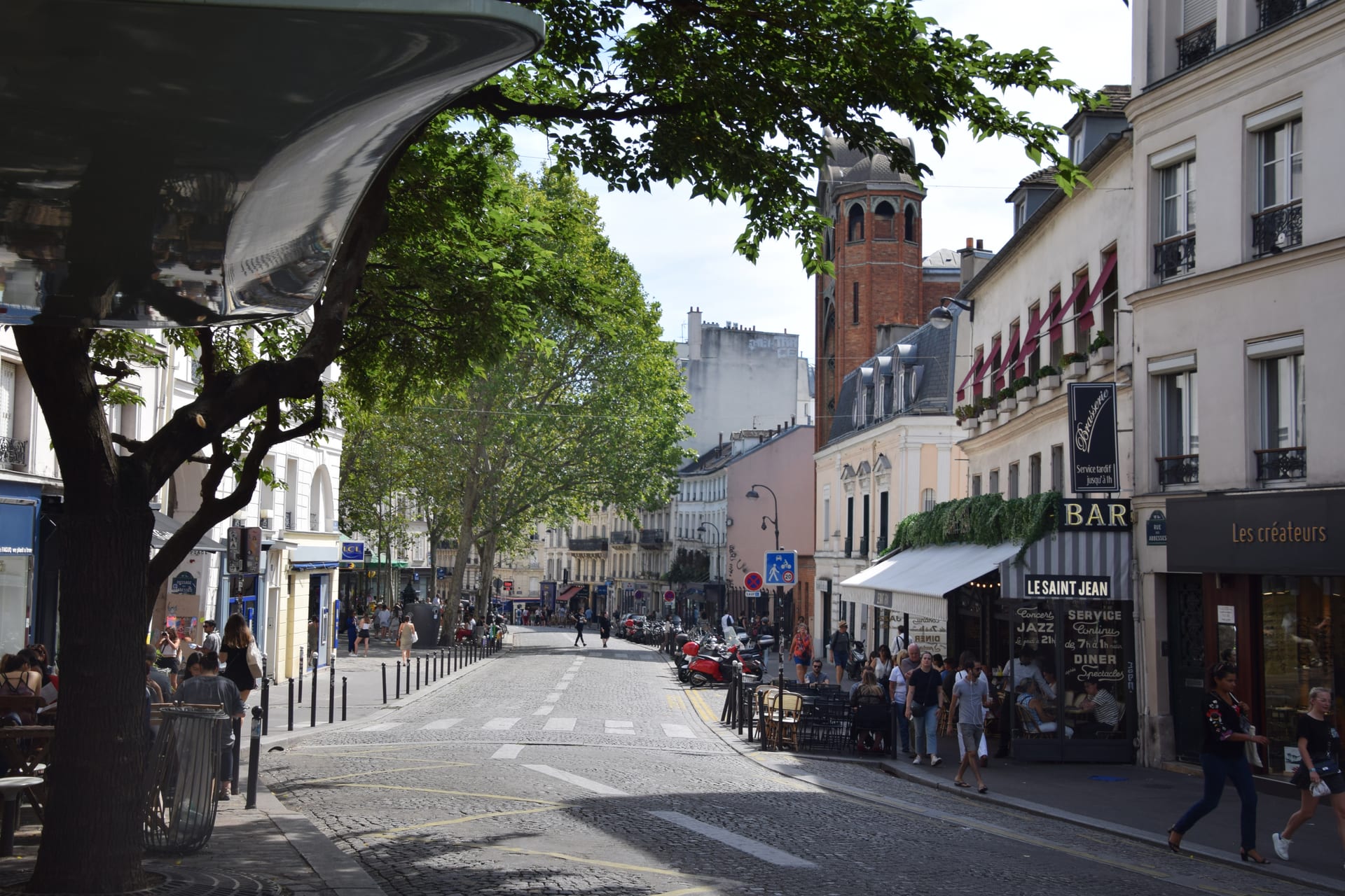 rue des abbesses a montmartre, una strada pedonale con caffe e il bar le saint jean, la torre campanaria in mattoni sullo sfondo e persone che passeggiano sotto gli alberi