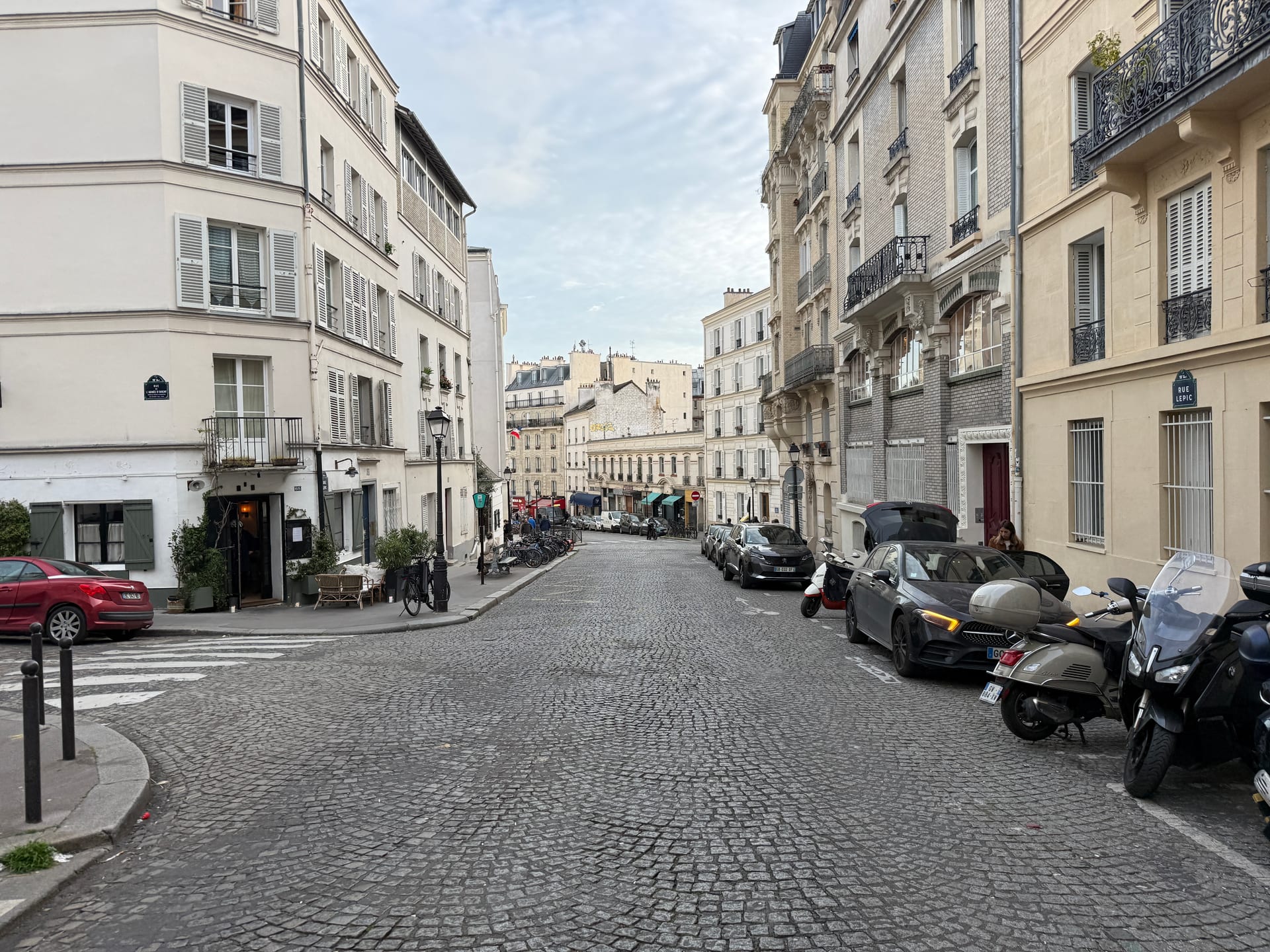 rue lepic a montmartre, una strada acciottolata in salita fiancheggiata da palazzi parigini, con auto e scooter parcheggiati sul lato destro