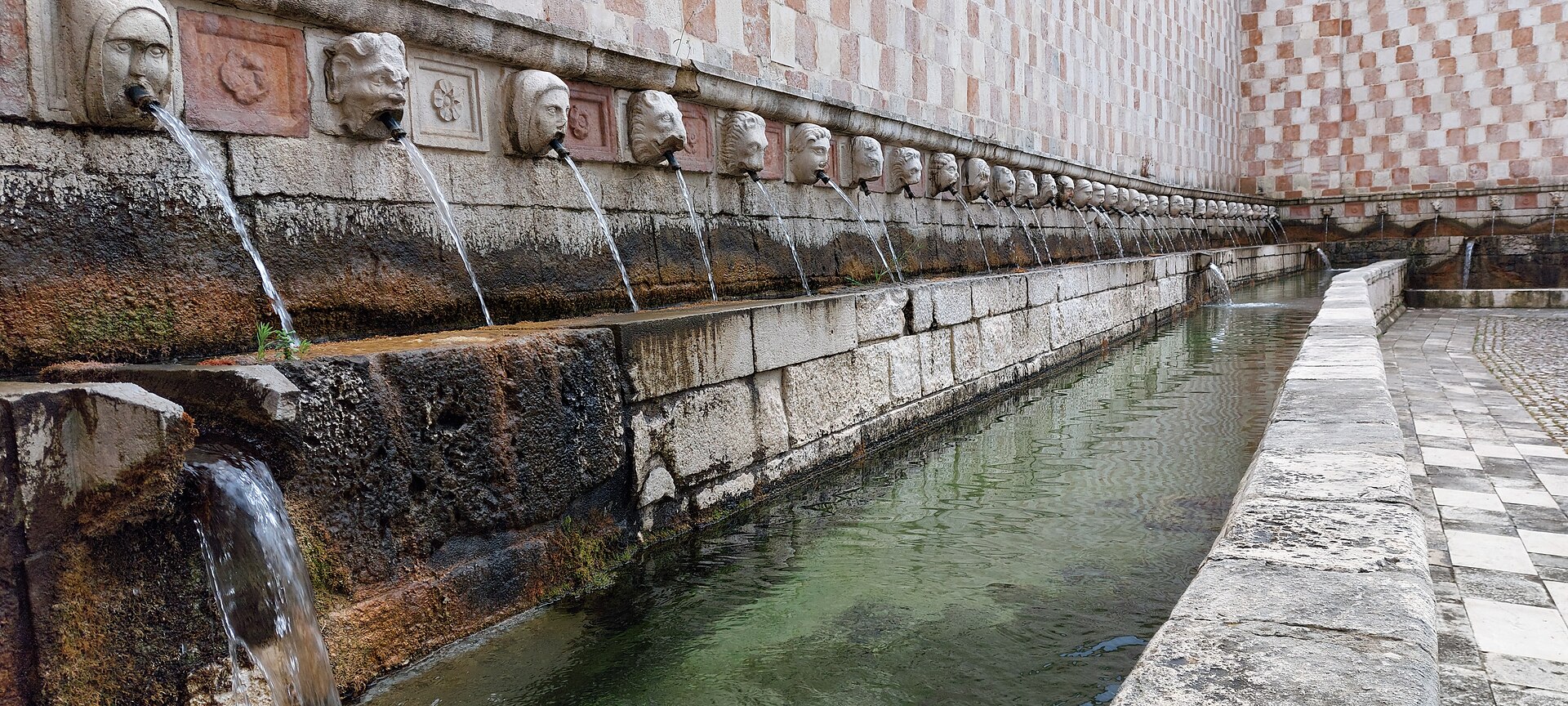 la fila in prospettiva dei mascheroni di pietra della fontana delle 99 cannelle con i getti d acqua nel canale