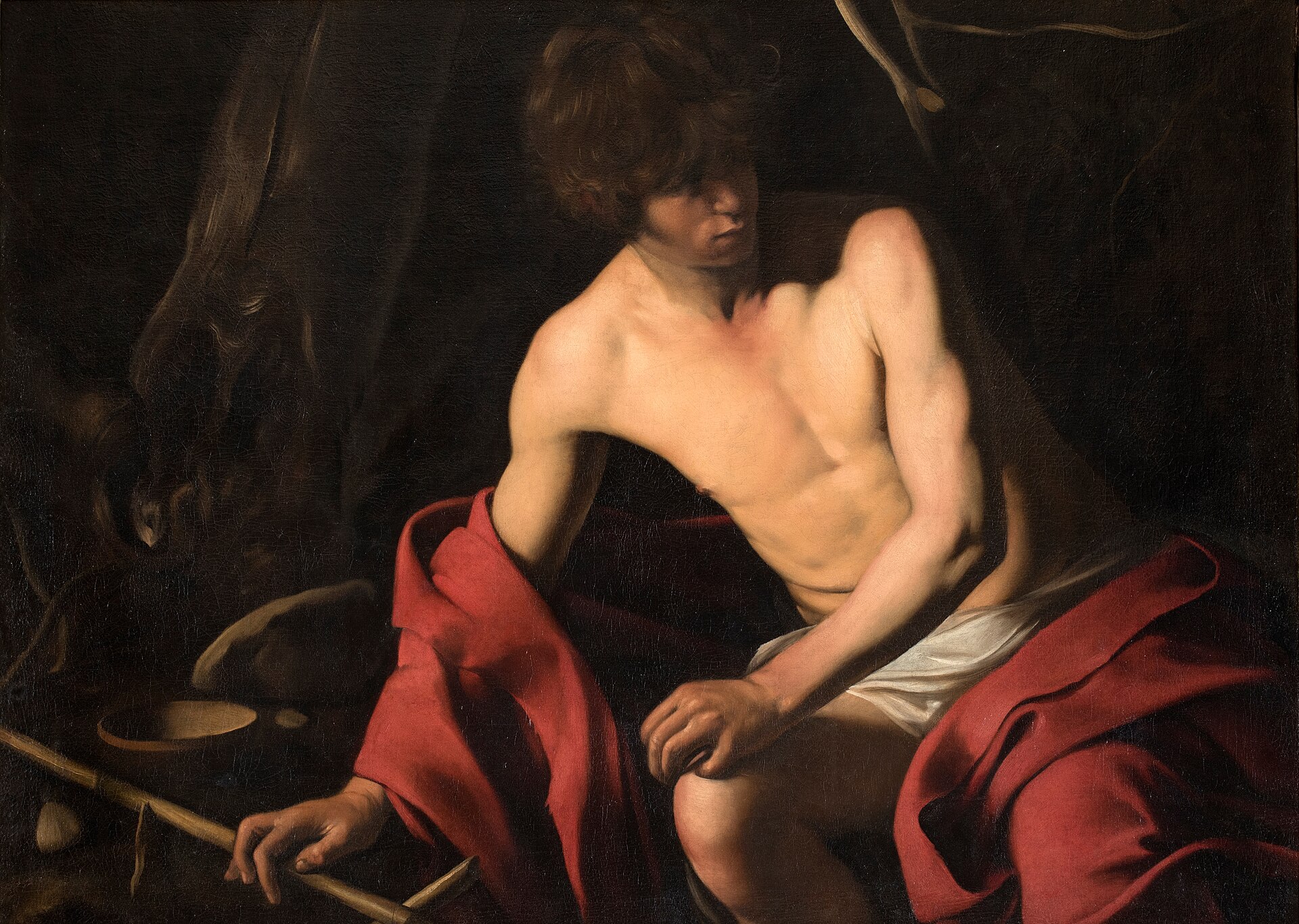 il san giovanni battista di caravaggio, un ragazzo seduto a torso nudo in un mantello rosso, con la mano sulla canna a forma di croce e una ciotola a terra, su un fondo scuro di tronchi