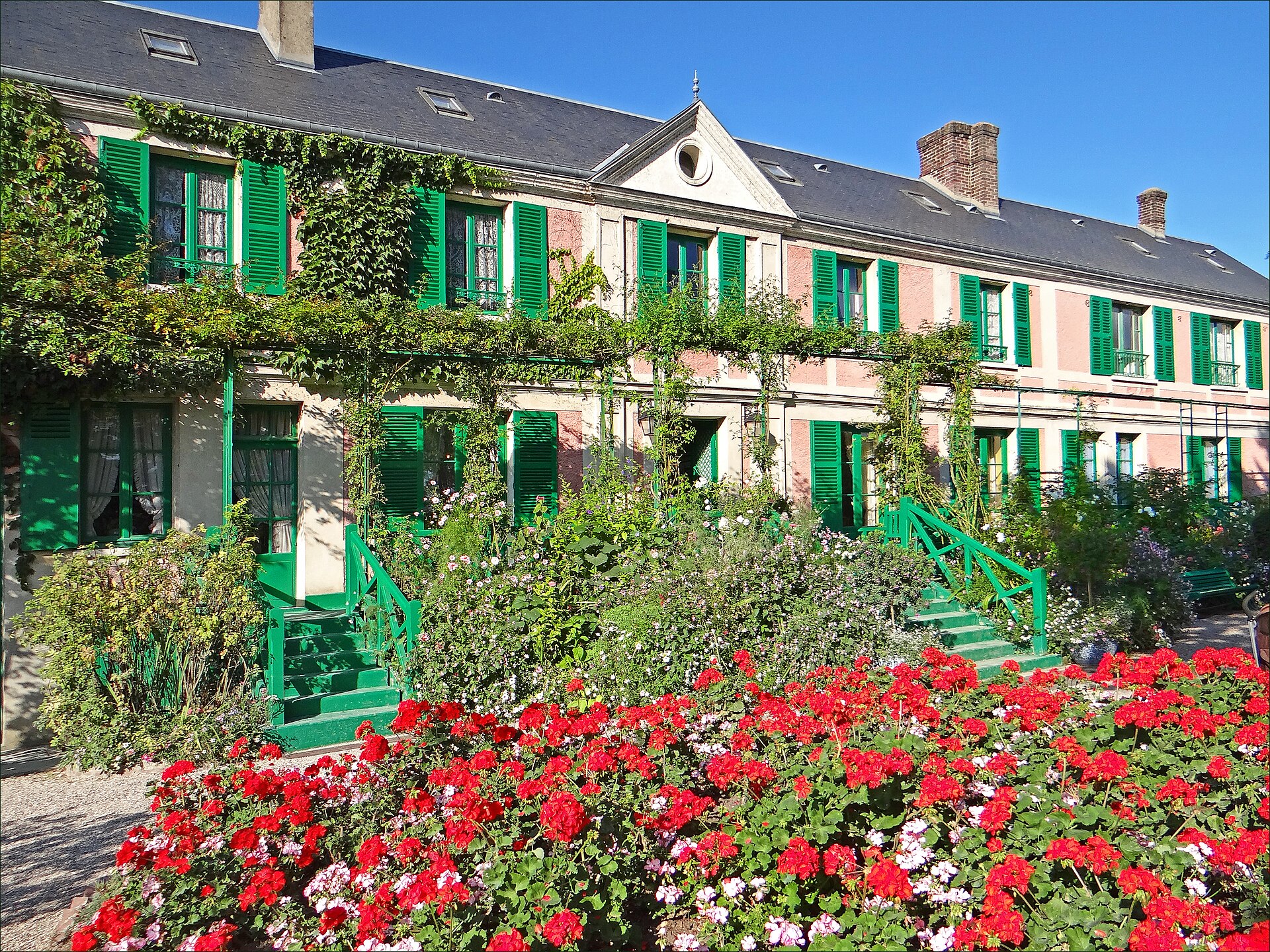 la facciata rosa della casa di claude monet a giverny, con le persiane verdi e le aiuole di gerani rossi in primo piano