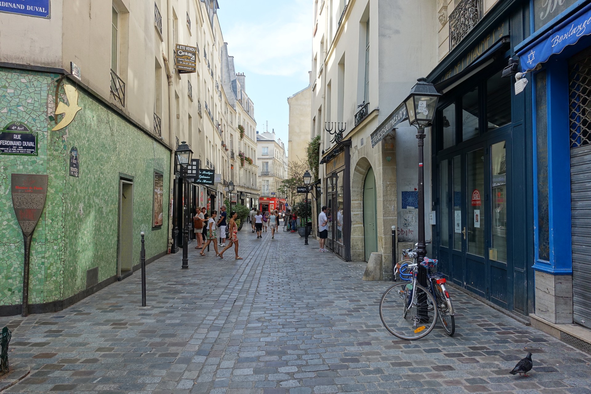 una strada del marais a parigi con un muro a mosaico verde, un insegna rue des rosiers e persone che passeggiano sul selciato
