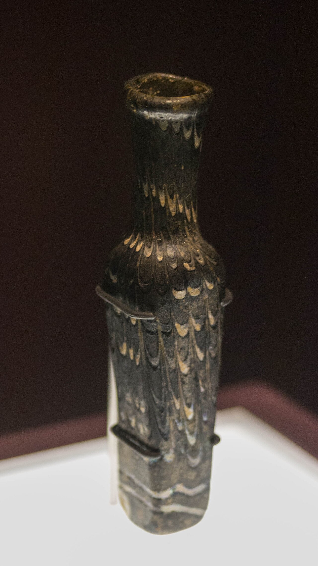 piccola fiala da profumo in vetro scuro con il decoro chiaro a piume, il collo stretto e la base quadrata, su un piedistallo bianco