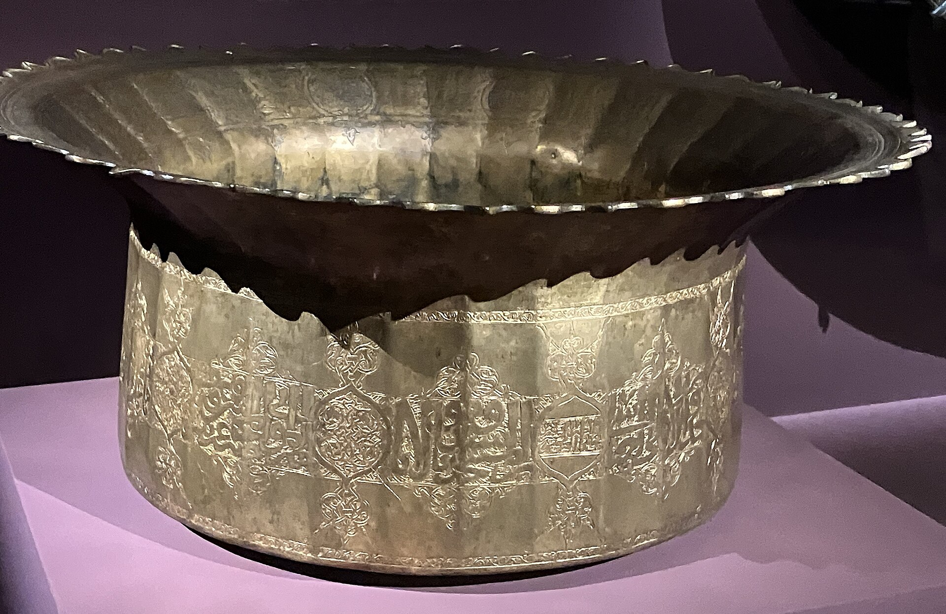 grande bacile in lega di rame dal bordo svasato e dentellato, con una fascia di cartigli e iscrizioni incise sul corpo cilindrico, su un piedistallo illuminato di lilla