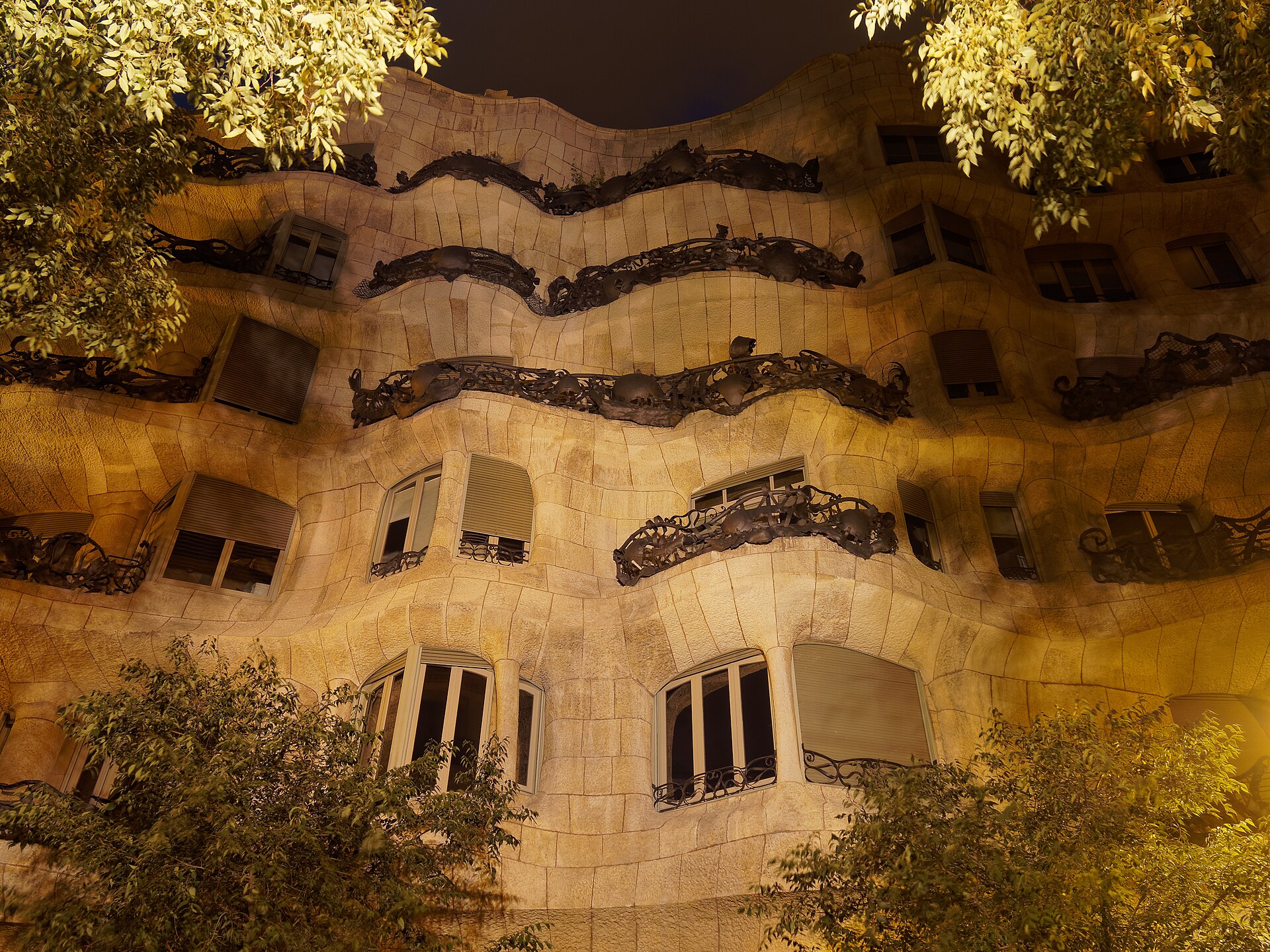 la facciata ondulata di pietra della pedrera illuminata la sera, con i balconi in ferro battuto e gli alberi davanti