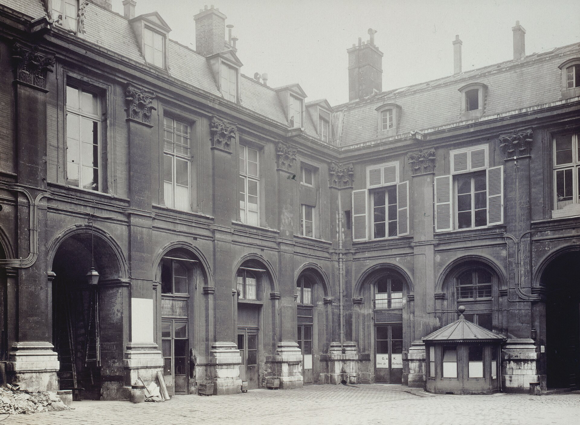 il cortile dell hotel de saint aignan fotografato nel 1916, in bianco e nero, con le botteghe sotto le arcate, le insegne dipinte e un chiosco di legno al centro