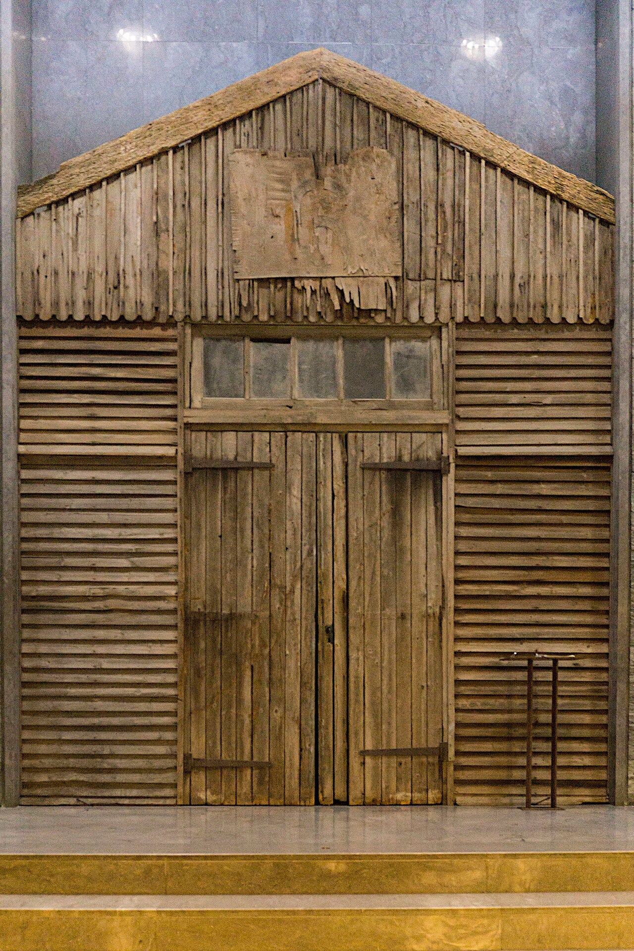 la facciata a timpano di una baracca di legno del campo di internamento di beaune la rolande rimontata dentro il museo contro una parete di marmo grigio, con le assi grezze, la doppia porta centrale, una fascia di vetri sopra la porta e un cartello lacero nel timpano