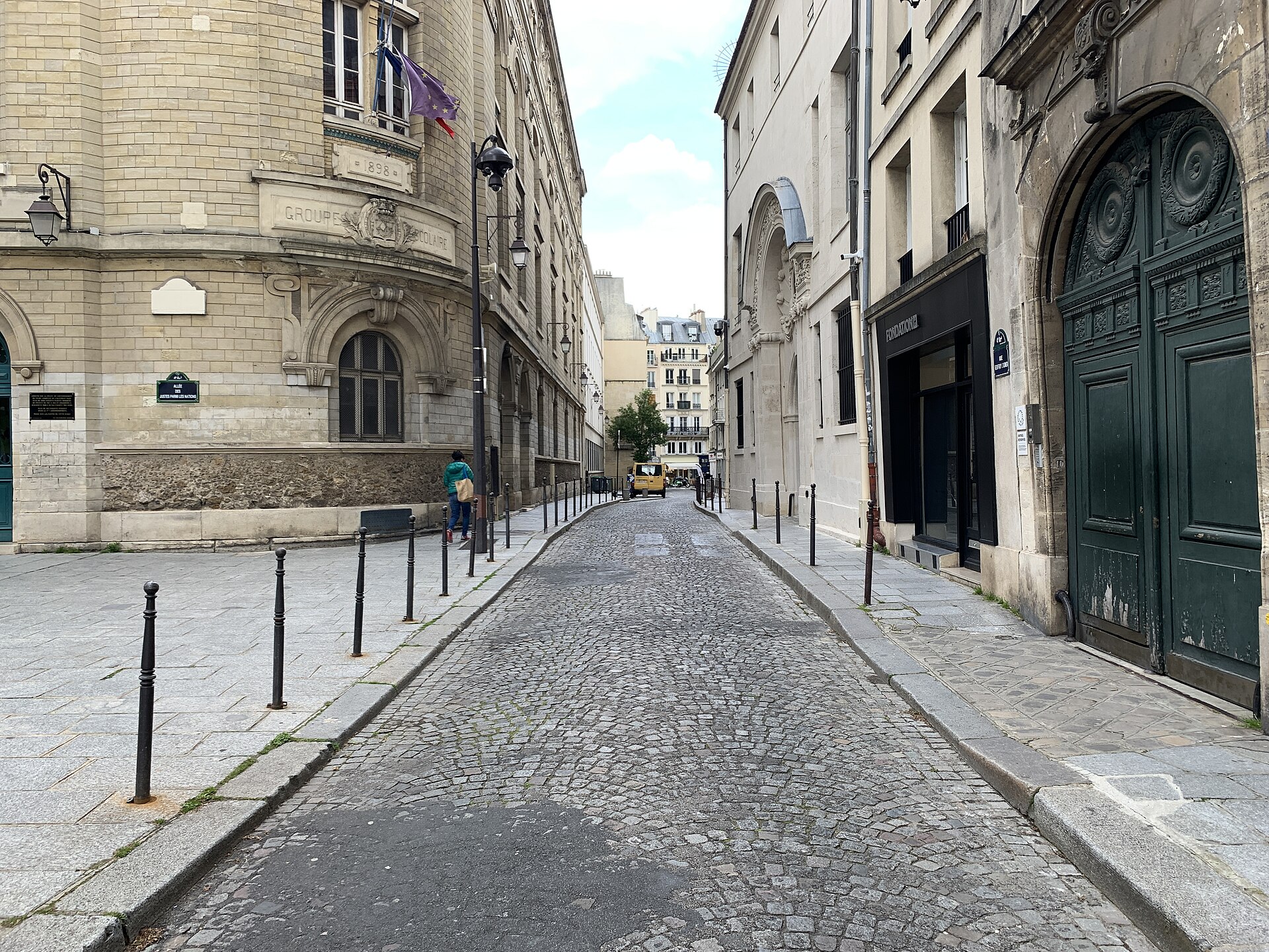 la rue geoffroy l asnier vista da terra, selciato di porfido fra due file di paracarri, a sinistra l angolo di un edificio scolastico del 1898 con la targa alle des justes parmi les nations e la bandiera francese, a destra i portoni verdi degli hotel particuliers del marais