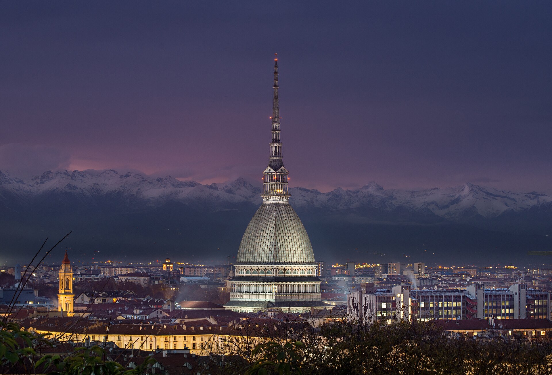 la mole antonelliana iluminada al anochecer con los alpes nevados detras y las luces de turin