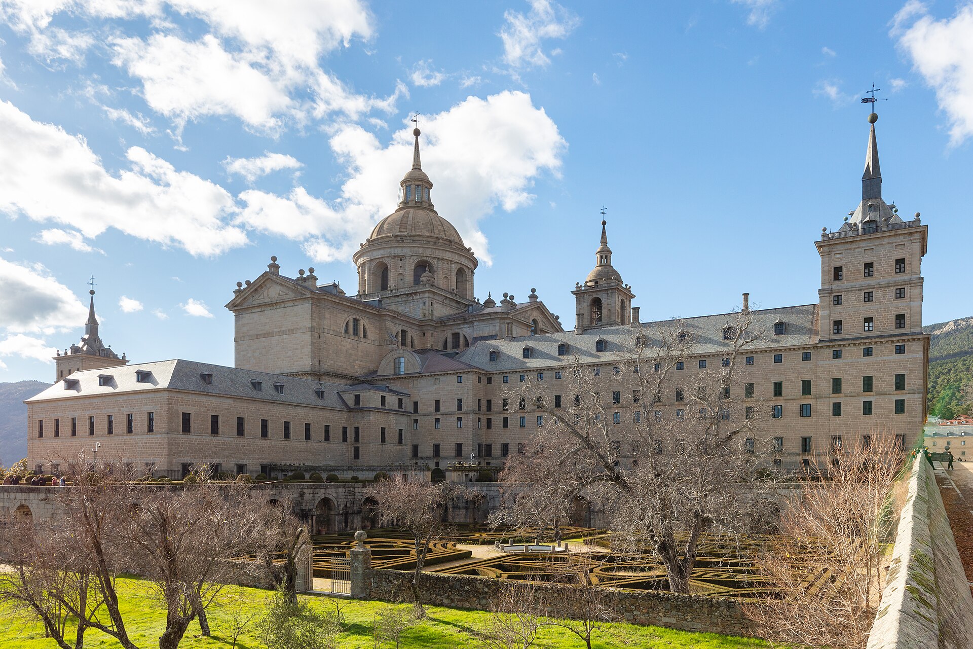 il monastero dell’escorial visto dal giardino dei frati, un enorme edificio di granito grigio con la cupola della basilica, le torri angolari e i tetti di ardesia, il giardino a siepi basse davanti e un prato con alberi spogli in primo piano