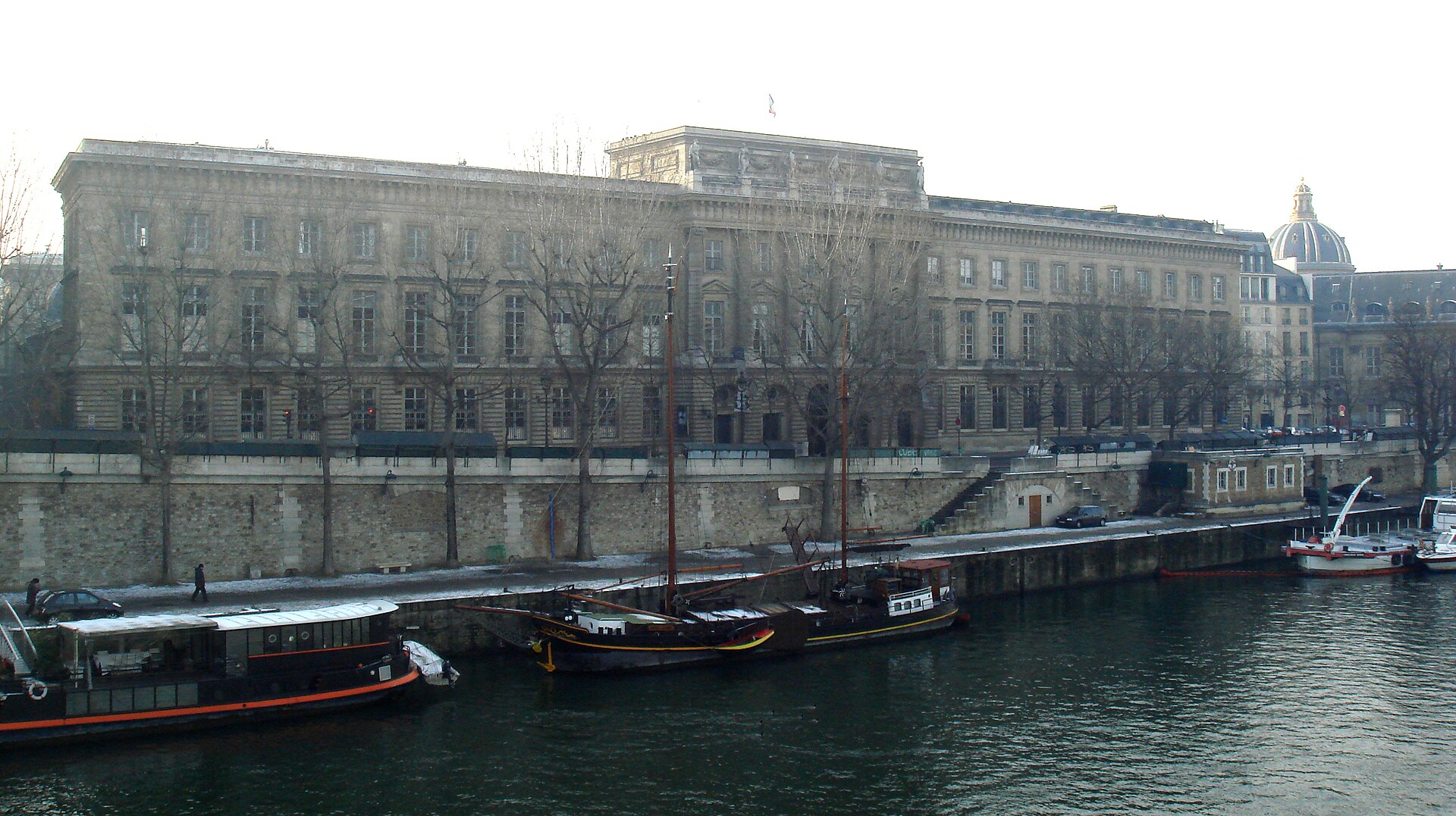 la lunga facciata neoclassica dell hotel de la monnaie vista dall altra riva della senna in inverno, con gli alberi spogli, le chiatte ormeggiate e la cupola dell institut de france sulla destra