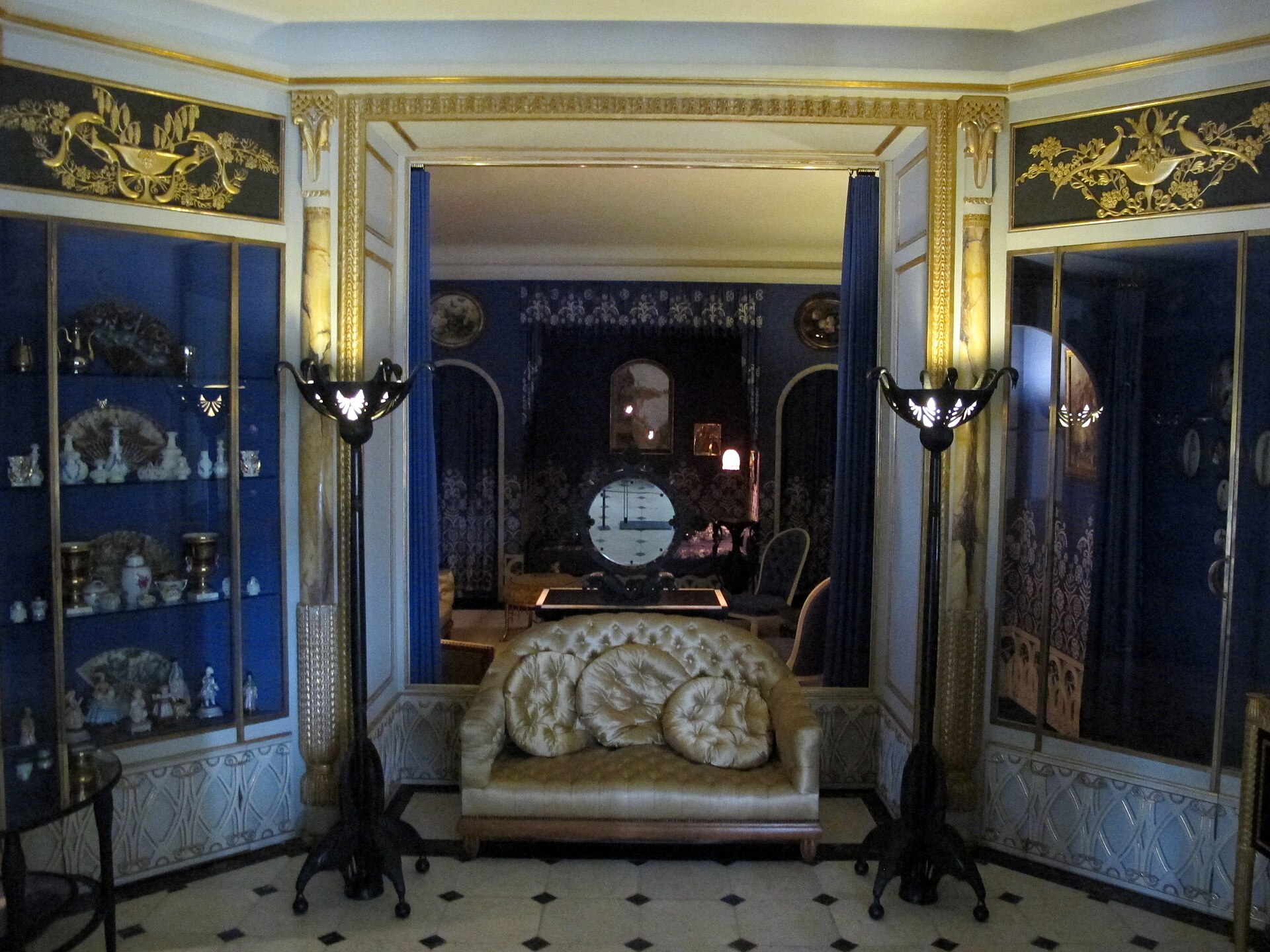 le boudoir de l appartement de jeanne lanvin au musee des arts decoratifs, avec les lambris gris et or, les reliefs dores aux oiseaux, deux lampadaires noirs, un canape creme et au-dela de la baie vitree la chambre bleue