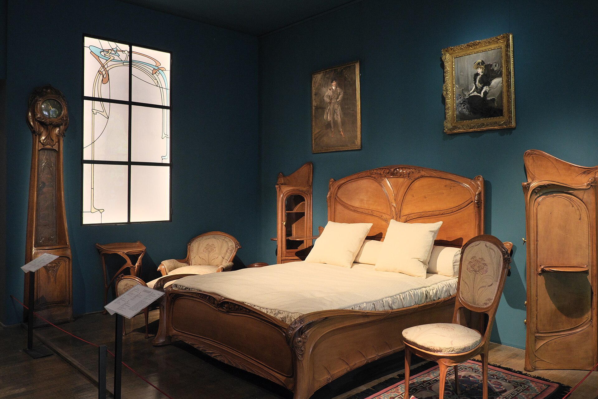 une chambre d hector guimard au musee des arts decoratifs, avec le lit en bois a la tete courbe, une horloge haute, les chaises et une fenetre aux vitrages sinueux sur des murs bleu canard