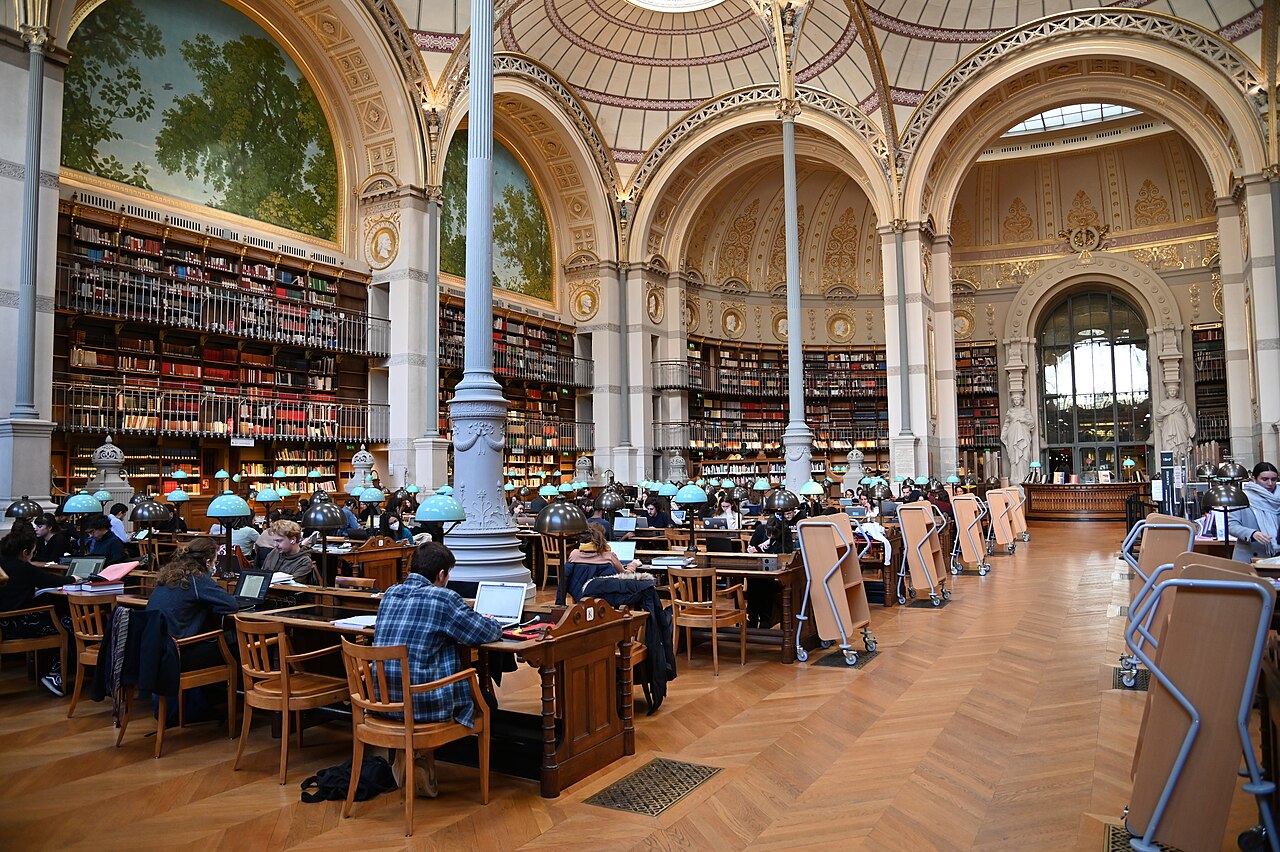 la salle labrouste con le cupole a lucernario sorrette da sottili colonne di ghisa, i paesaggi dipinti nelle lunette sopra gli scaffali e i tavoli di lettura con le lampade dal paralume verde