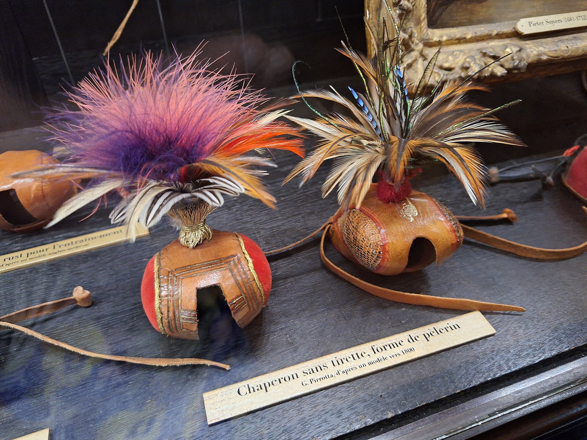 due cappucci da falconeria in una vetrina del musee de la chasse et de la nature, di cuoio rosso e marrone con ricami dorati e ciuffi di piume colorate, con i cartellini scritti a mano