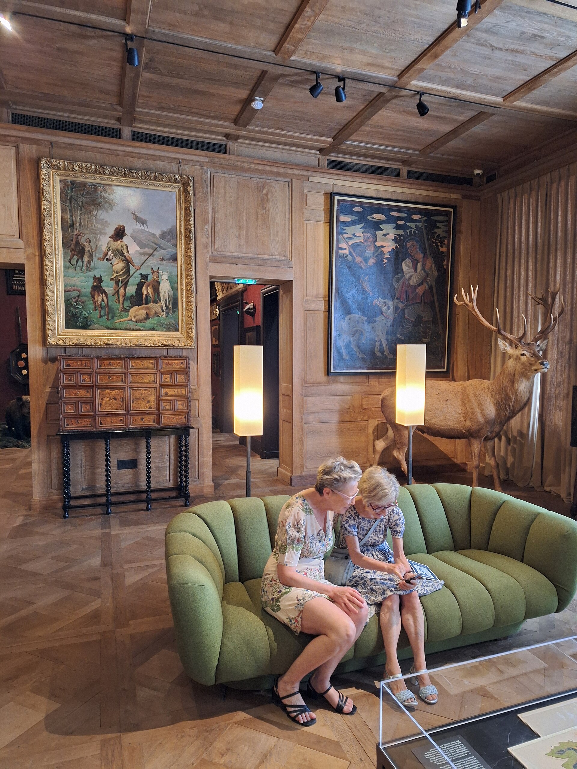 una sala del musee de la chasse et de la nature con le pareti di legno chiaro, due grandi dipinti di caccia, un mobile intarsiato, un divano verde e un cervo naturalizzato accanto alla tenda