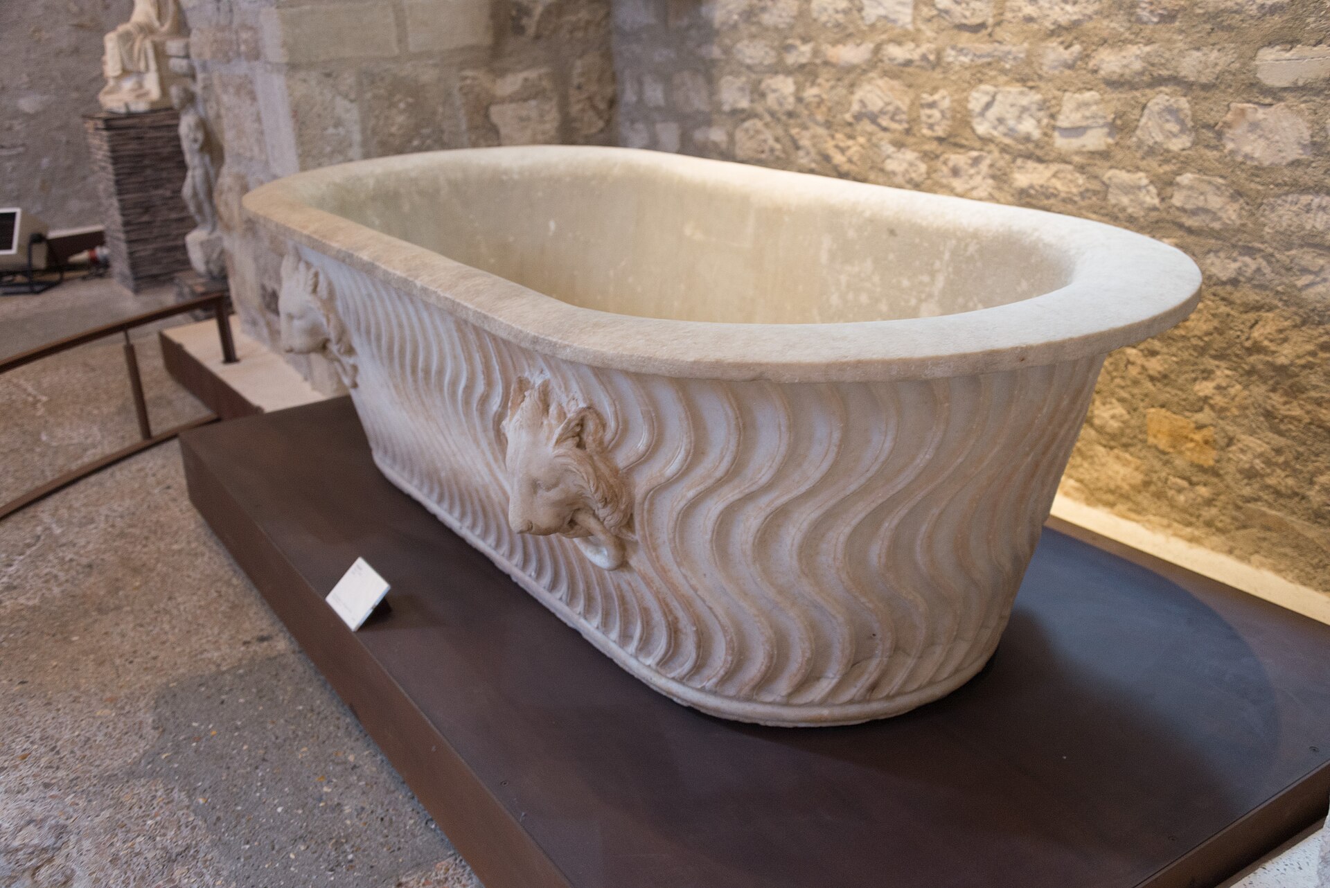 a roman white marble bath at the musee de cluny, with wave carved sides and a lion head in relief, in front of a stone wall