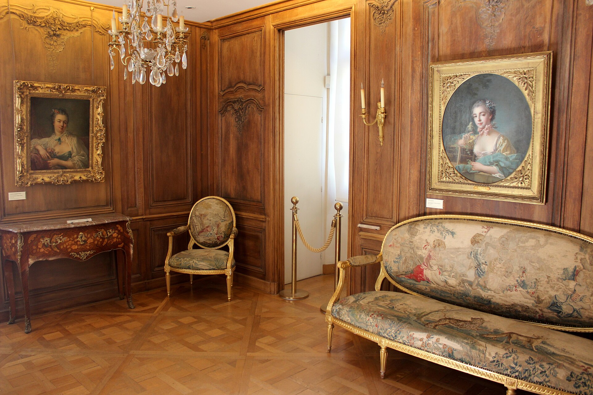 una sala del musee cognacq jay con le boiserie di quercia, due ritratti femminili settecenteschi incorniciati d oro, un divano coperto di arazzo e un lampadario di cristallo