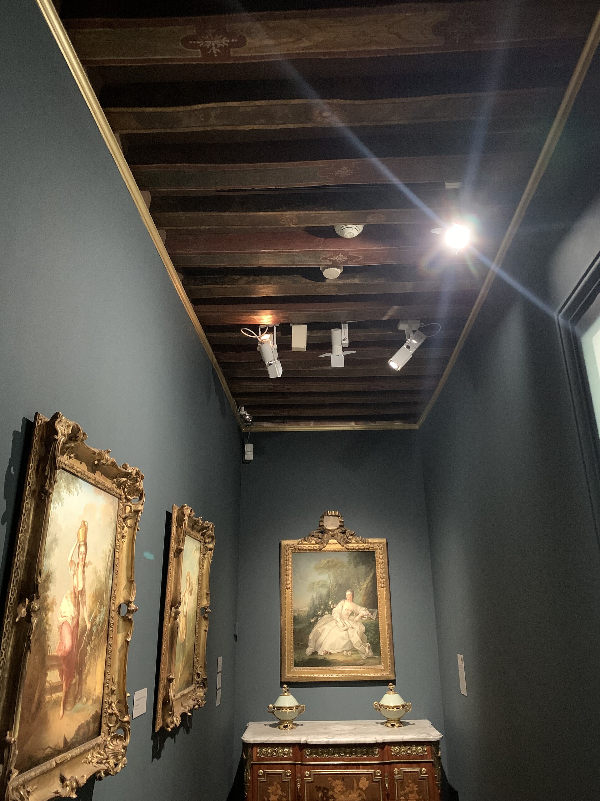 una sala del musee cognacq jay dalle pareti grigio azzurre, con due dipinti settecenteschi in cornice dorata, un comò intarsiato e il soffitto a travi di legno dipinte