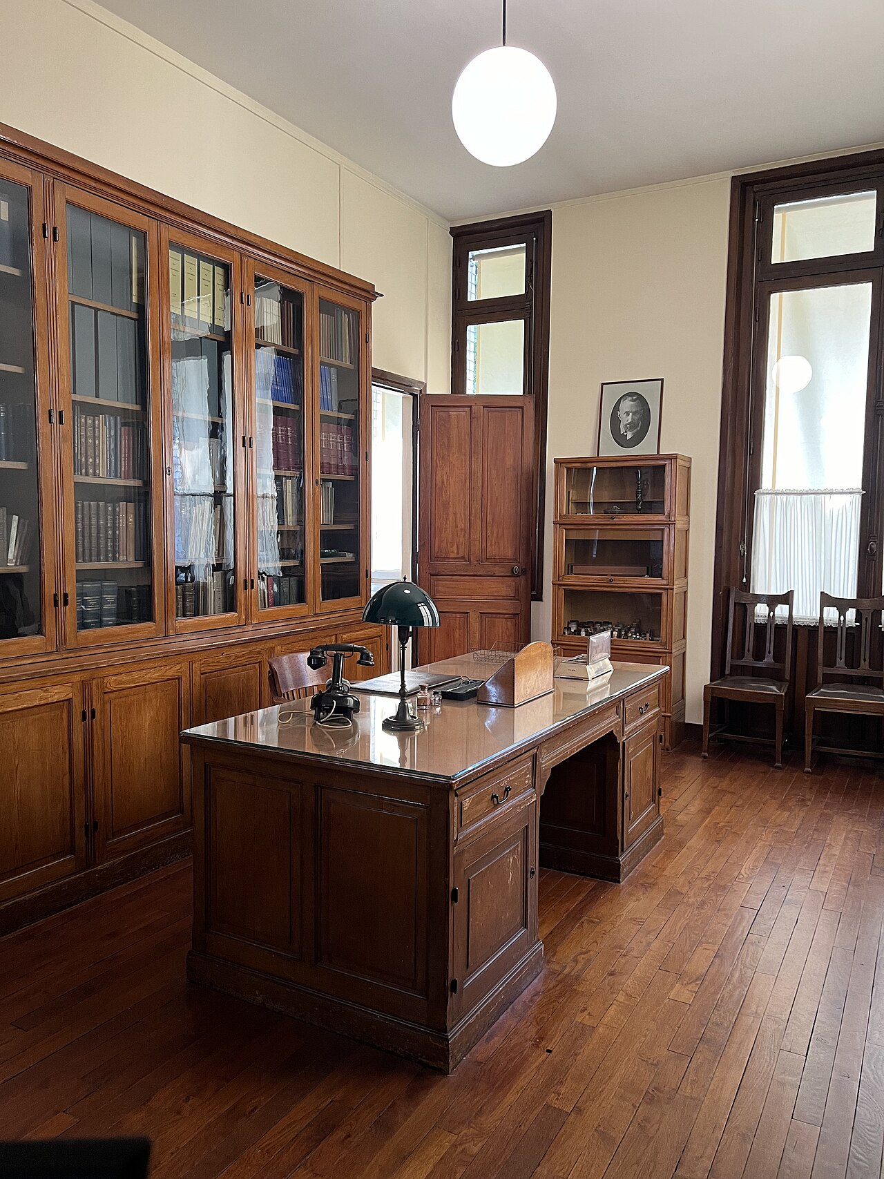 l ufficio di marie curie con la grande scrivania di legno al centro, la lampada dal paralume verde e il telefono nero a disco, la libreria a vetri lungo la parete e un ritratto fotografico incorniciato su un mobiletto
