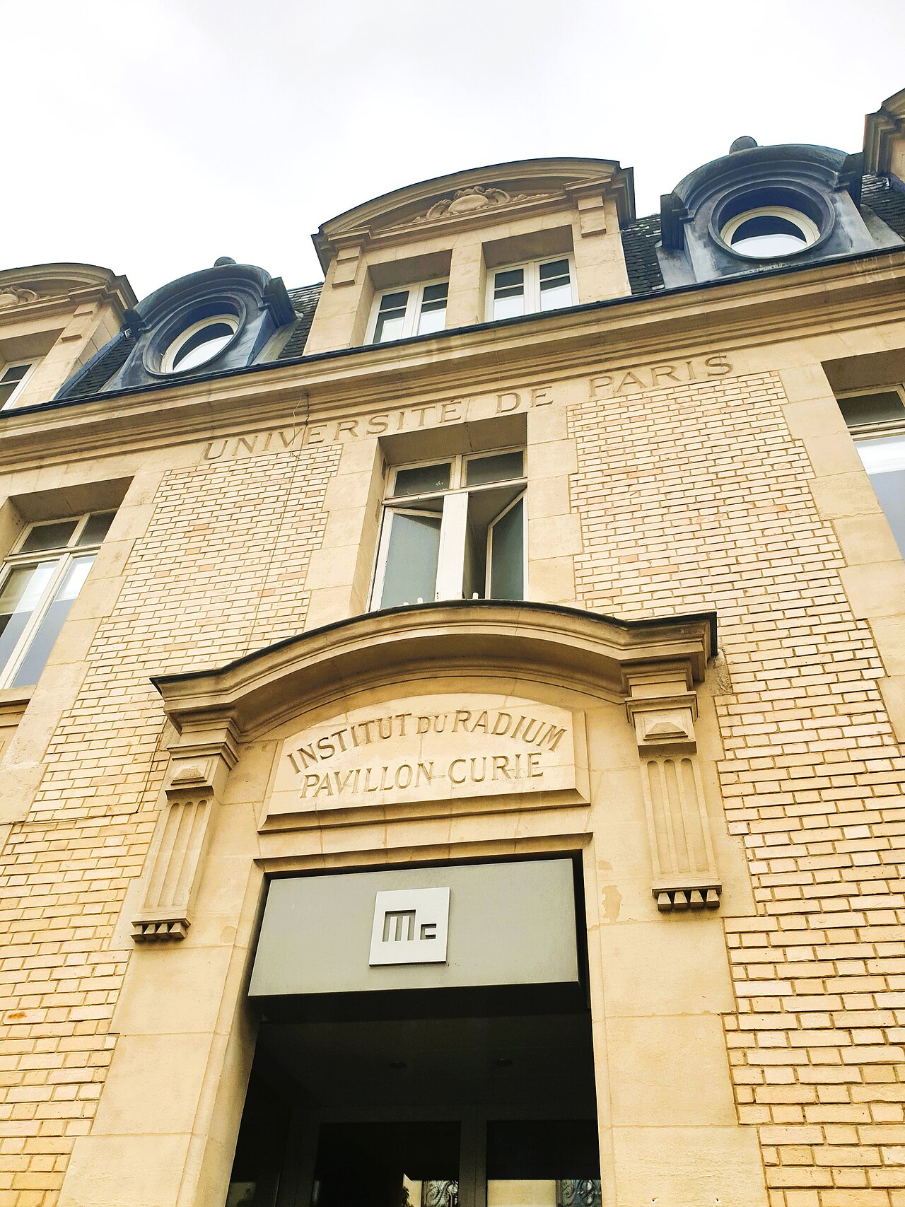 la facciata di mattoni chiari del pavillon curie vista dal basso, con la scritta scolpita universite de paris sotto il tetto e, sopra l ingresso, institut du radium pavillon curie