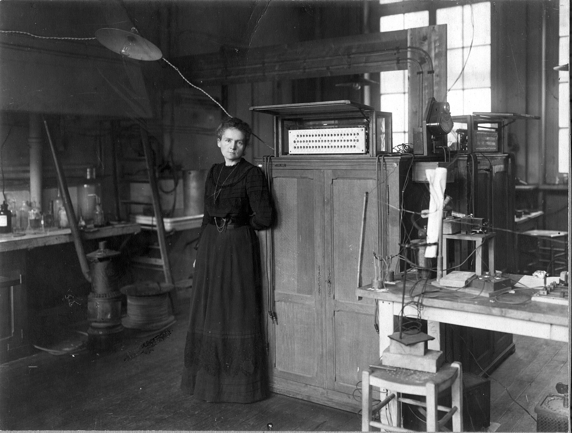 fotografia in bianco e nero del 1912: marie curie in piedi con un abito scuro lungo, appoggiata a un armadio sormontato da un quadro di connessioni elettriche, in un laboratorio pieno di strumenti e cavi
