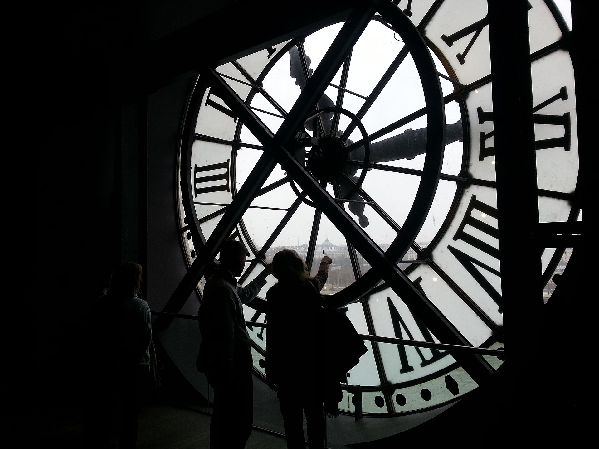 des visiteurs en contre-jour derriere la grande horloge du cinquieme etage du musee d orsay, qui regardent paris a travers le cadran transparent aux chiffres romains