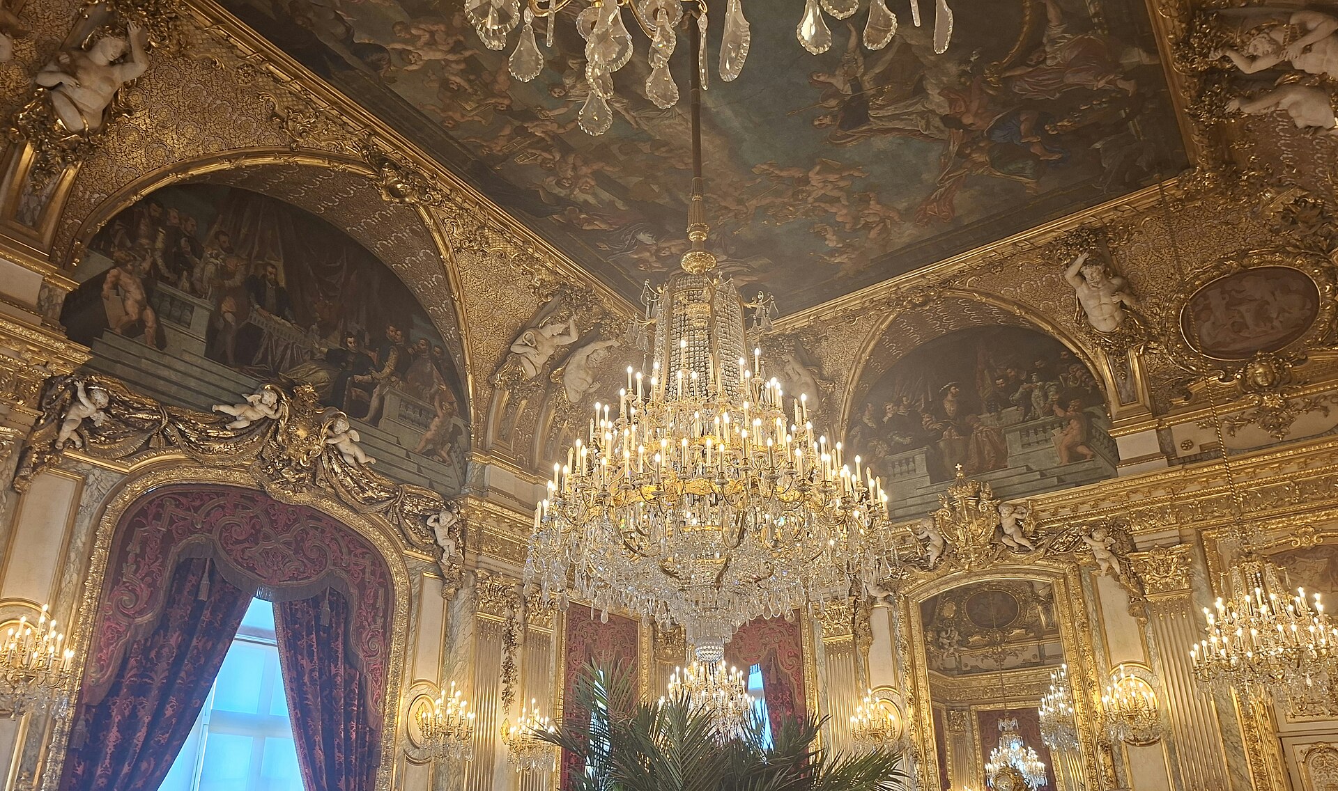 le grand salon des appartements napoleon iii au louvre, avec les lustres de cristal allumes, le plafond peint, les rideaux rouges et les canapes