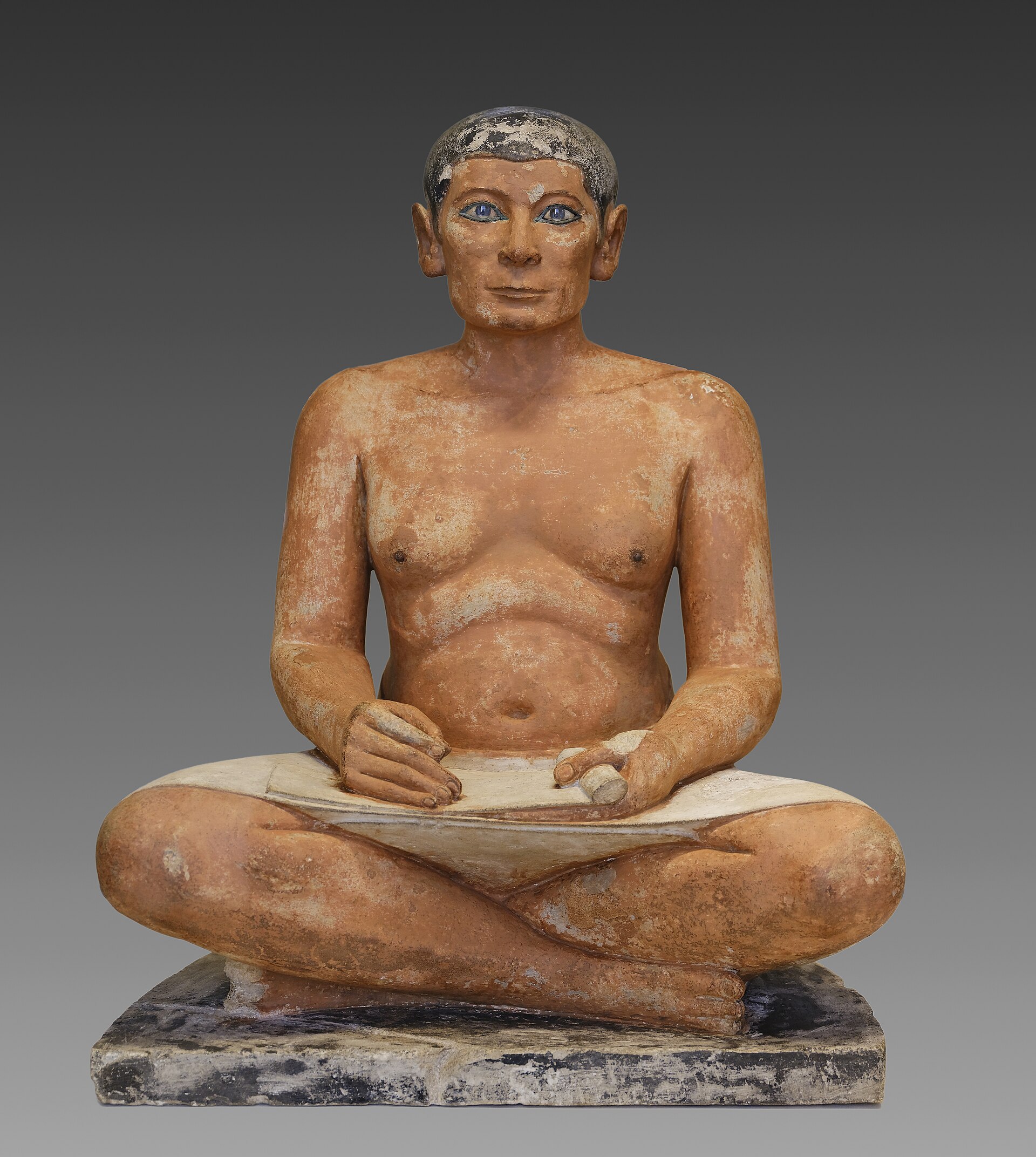 le scribe accroupi du louvre, un homme assis en tailleur avec le rouleau de papyrus ouvert sur les genoux, les cheveux ras et les yeux blancs et sombres qui regardent droit devant