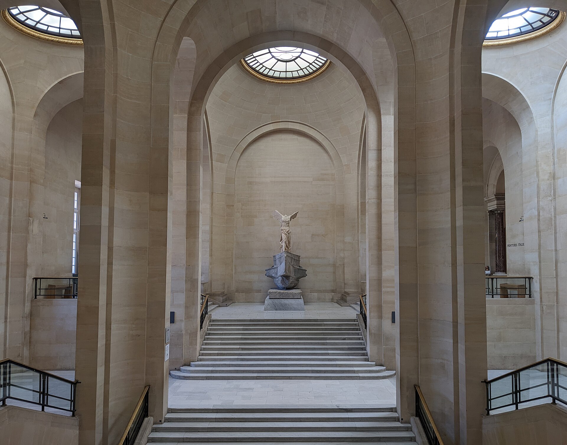 l escalier daru du louvre vu d en bas, avec la victoire de samothrace sur le palier en haut et des arcades de pierre claire de chaque cote