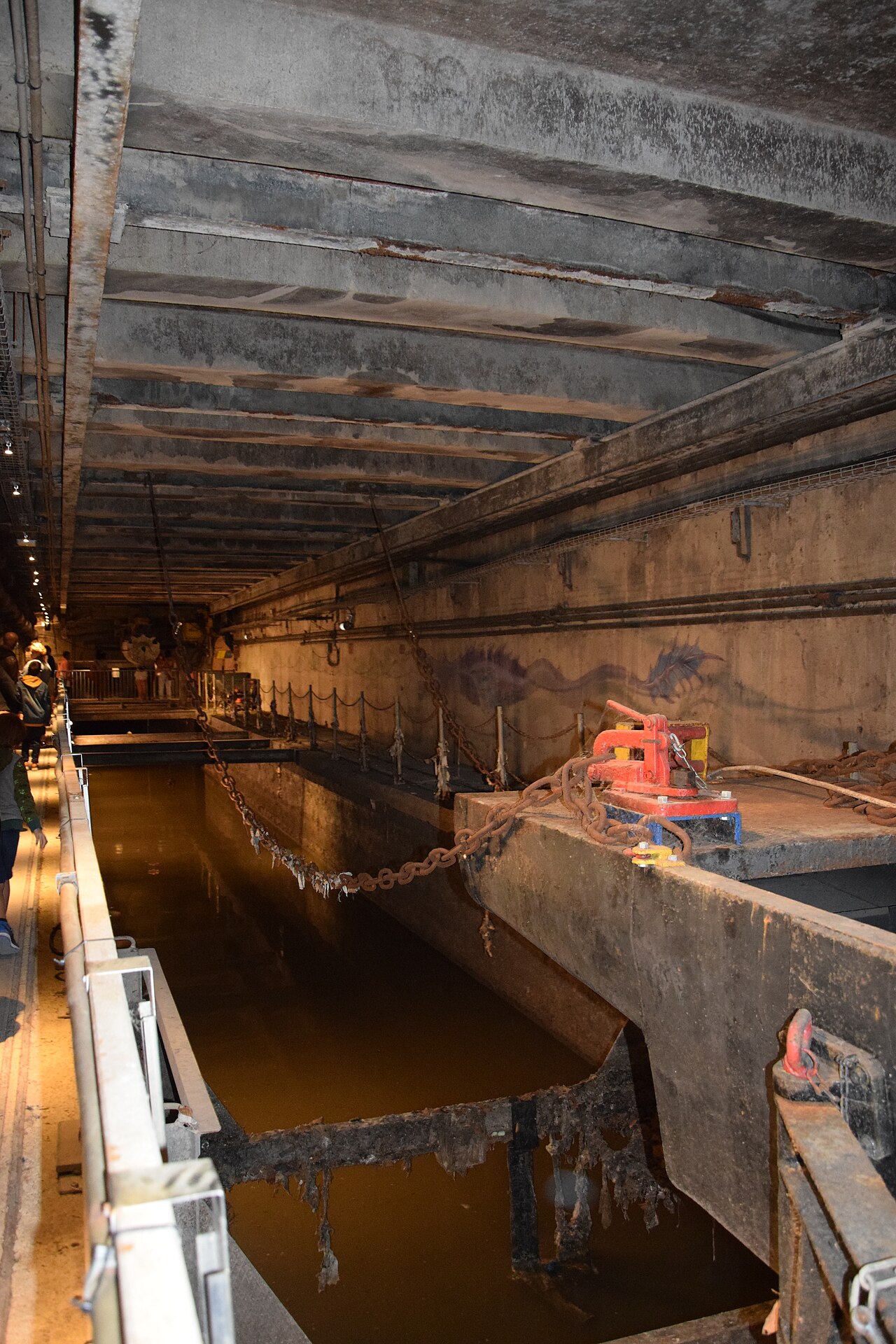 l interno di un grande collettore con il canale d acqua scura al centro, una macchina di spurgo arrugginita sospesa su rotaie con le sue catene, i tubi appesi alla volta e alcuni visitatori sulla passerella in fondo