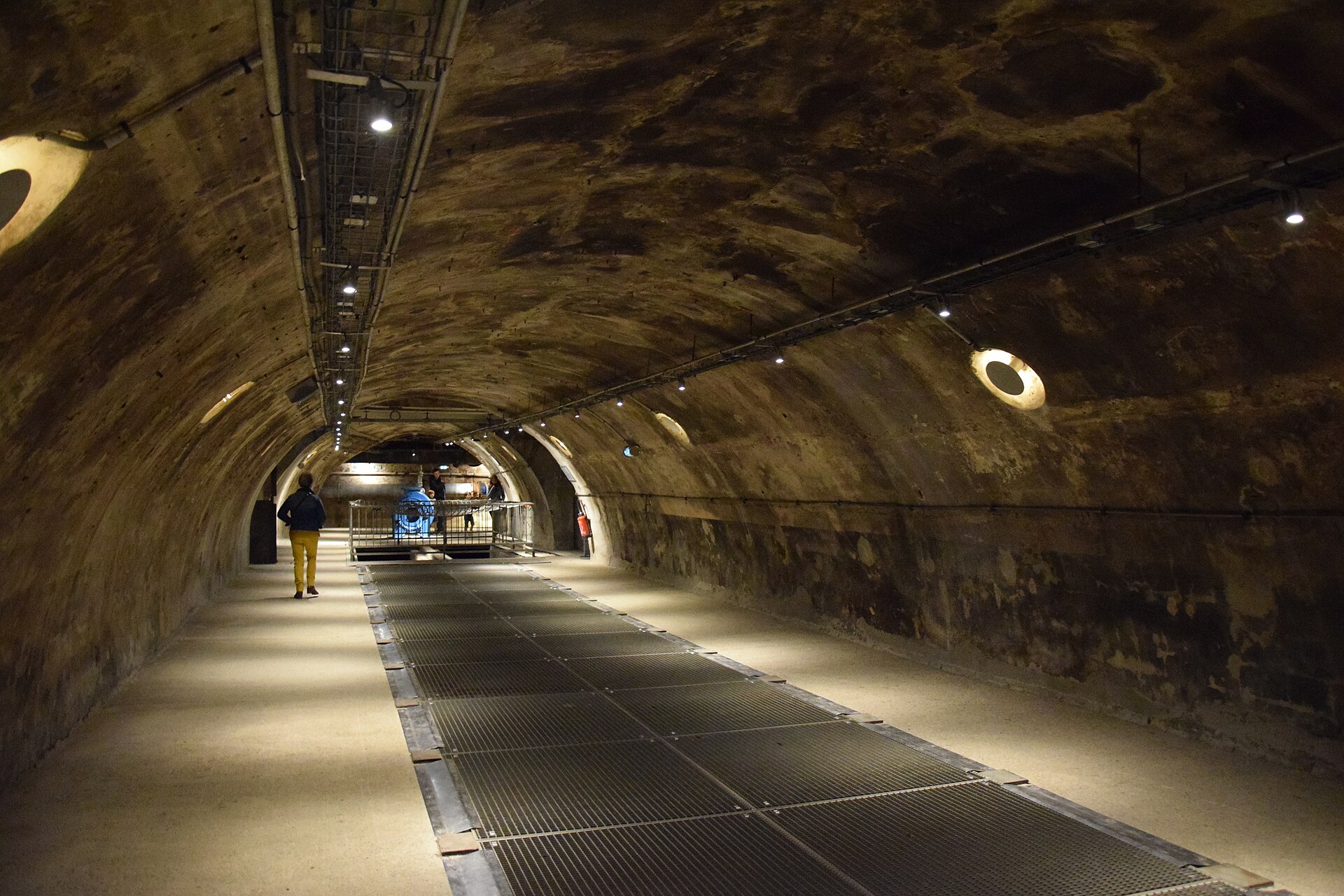 una grande galleria fognaria a volta, con la passerella di grigliato metallico che corre al centro sopra il canale, le lampade ovali incassate nella parete di destra, i cavi e i tubi appesi alla volta e due visitatori in fondo accanto a una macchina azzurra