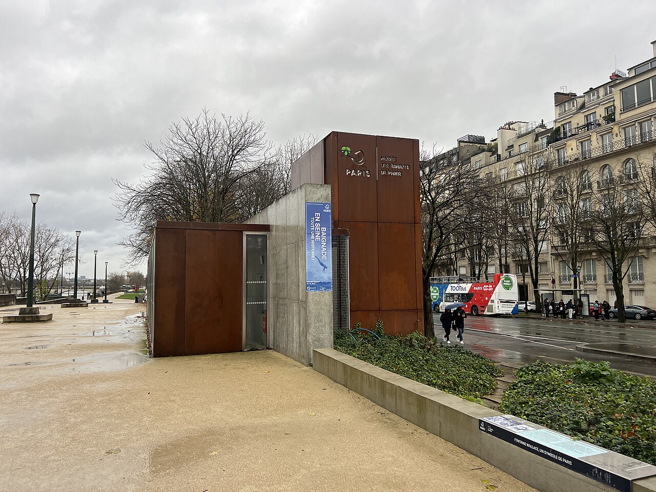 il padiglione d ingresso in acciaio corten sul lungosenna, con l insegna del museo e il logo della citta di parigi, i palazzi haussmanniani e il traffico sullo sfondo in una giornata di pioggia