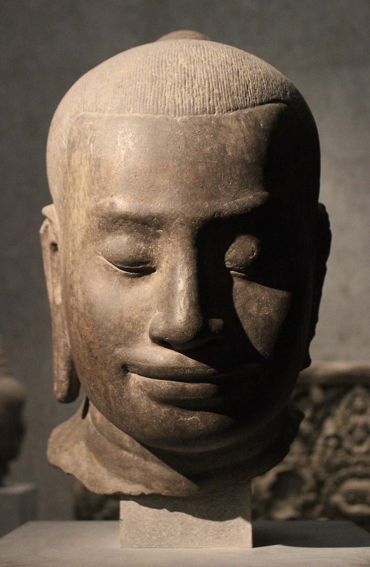 la testa in arenaria di jayavarman vii al musee guimet, il volto con gli occhi chiusi e un accenno di sorriso, i capelli a righe incise sottili e il collo spezzato
