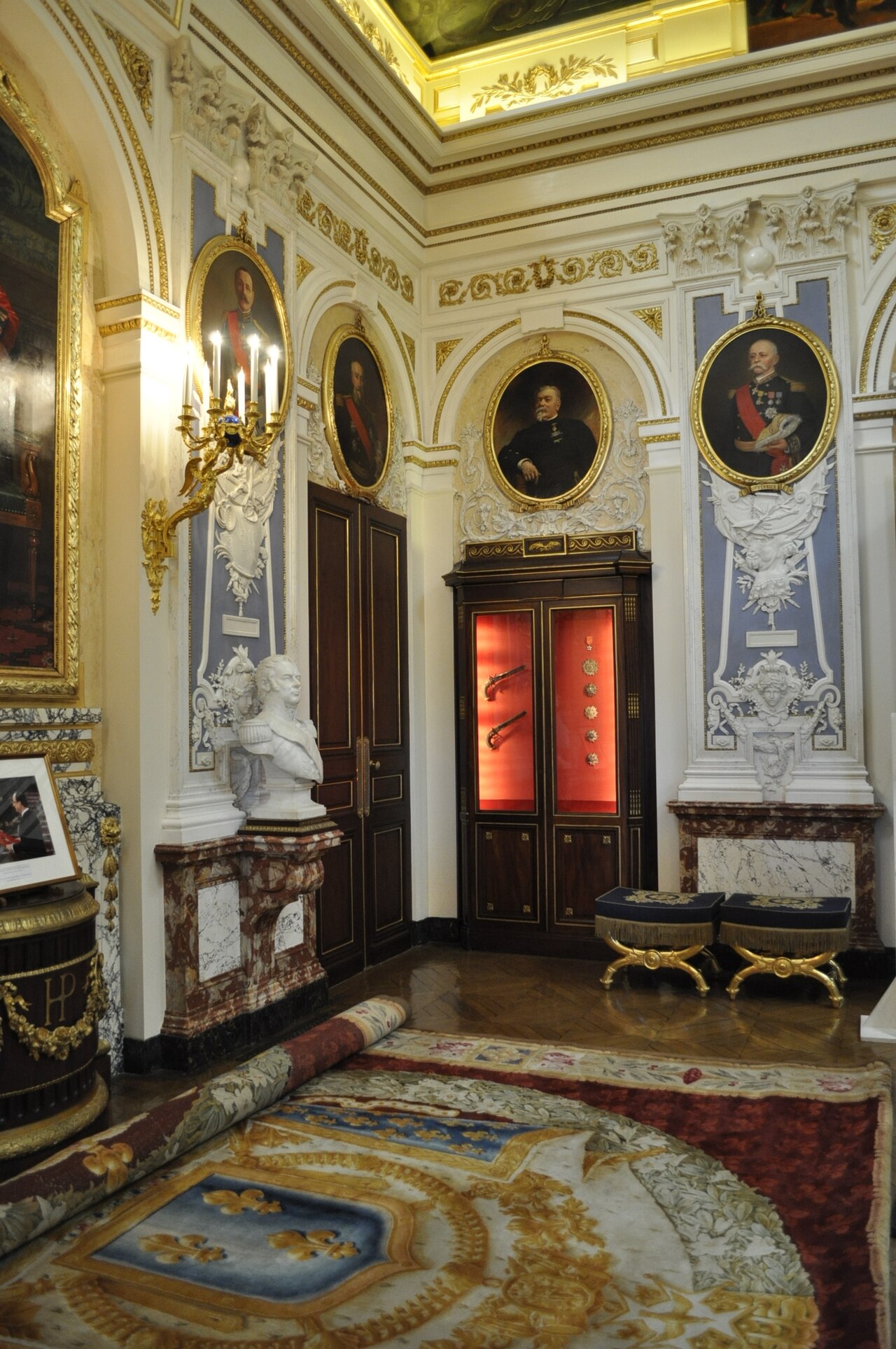 il salone dei grandi cancellieri con le pareti a boiserie bianca e oro, i ritratti ovali dei cancellieri nelle cornici dorate, un camino, un busto di marmo e una vetrina illuminata di rosso con pistole e decorazioni